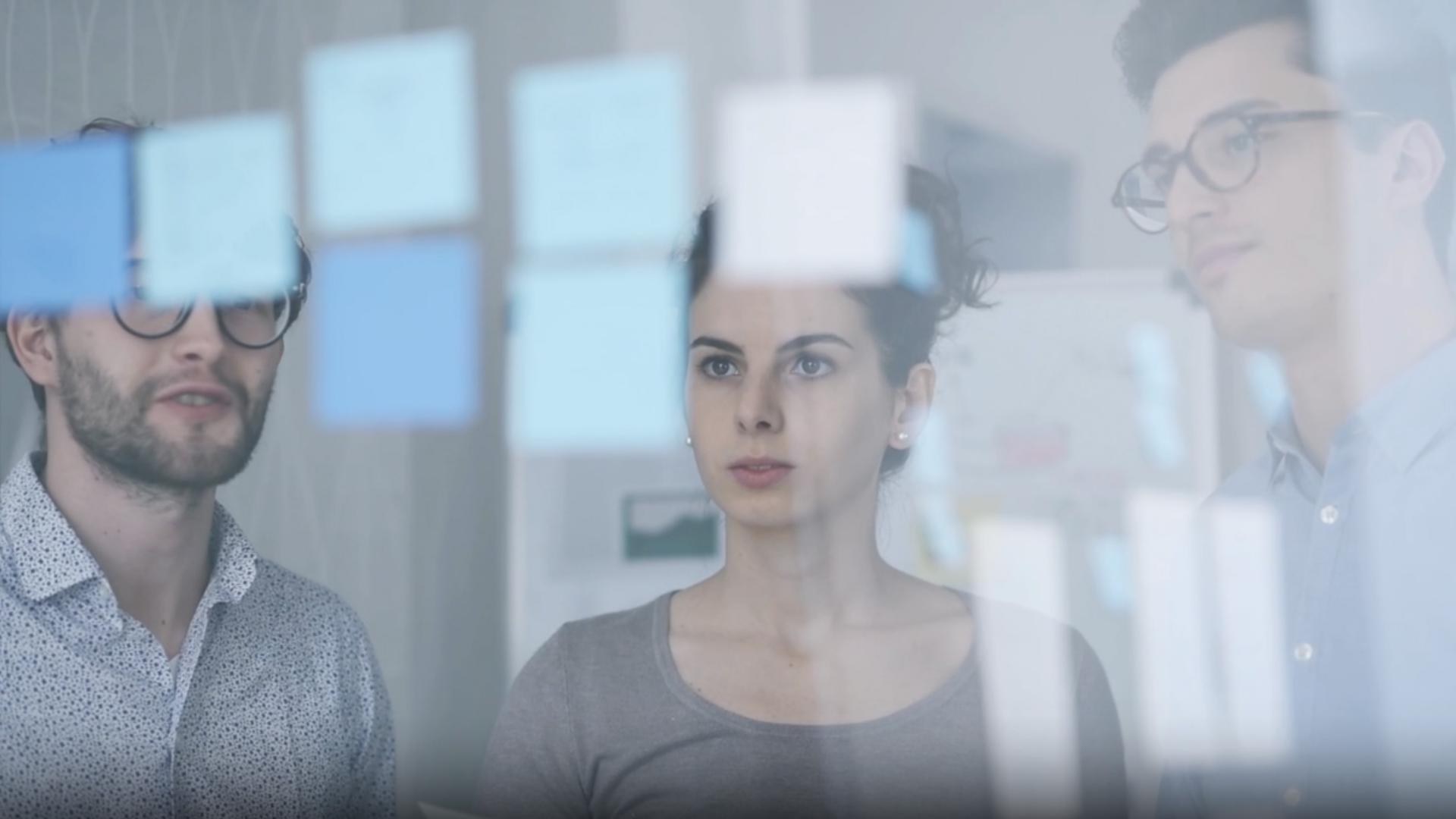 Three people are working on a transparent work wall that has many blue sticky notes attached to it.
