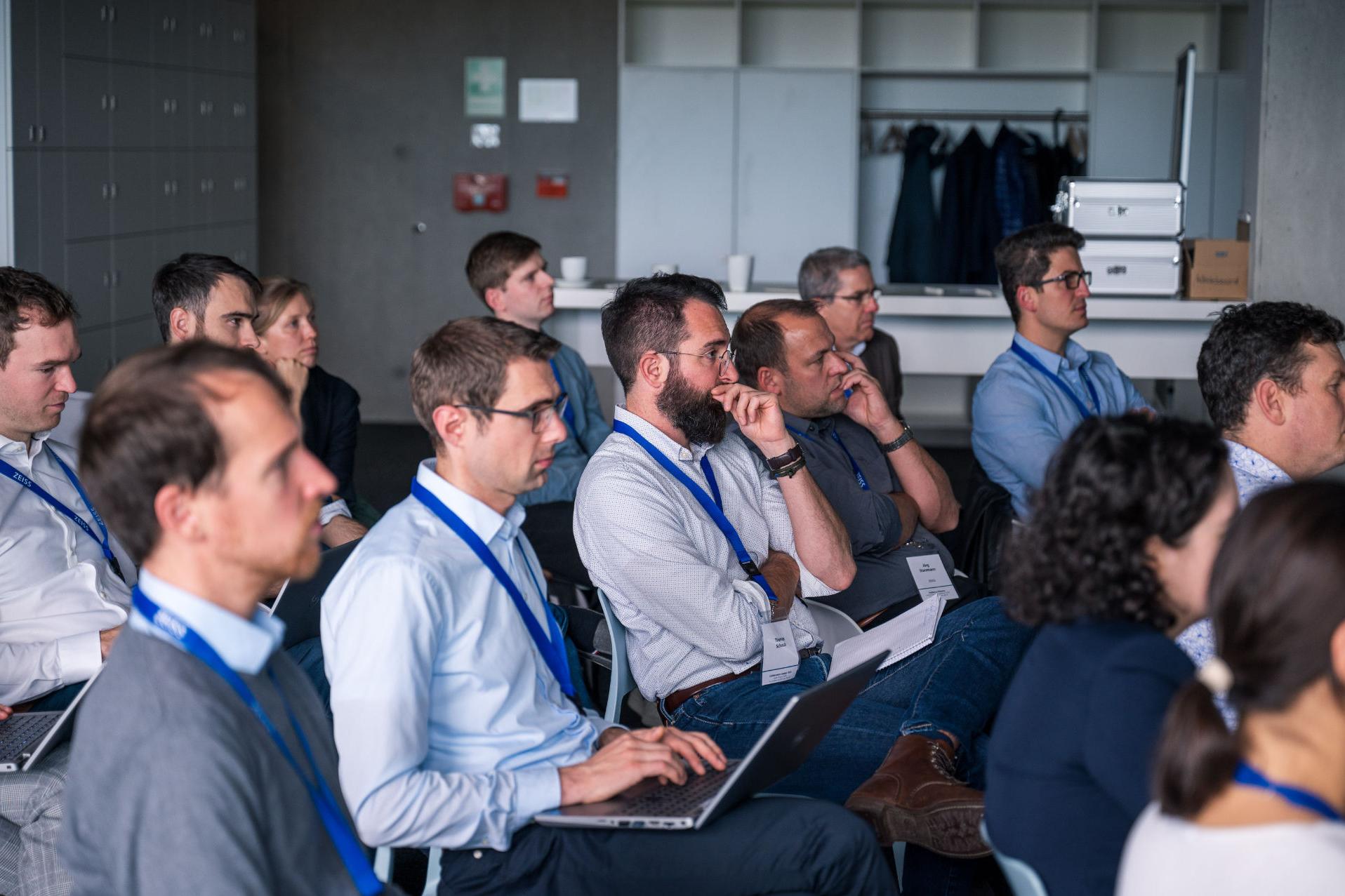 View of the audience made up mostly of men, attending a presentation. Most of the attendees are wearing white and blue button-down shirts and dark blue jeans.