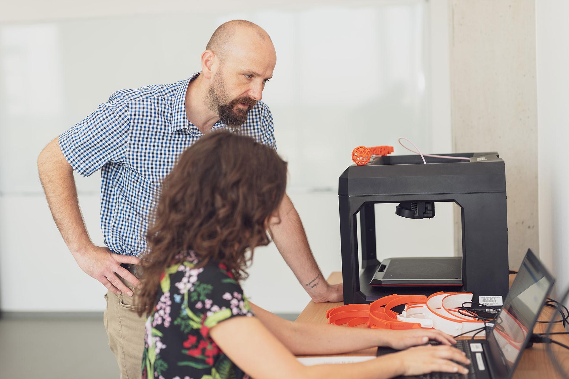 Two people are working in the Makerspace sitting at a desk using a laptop and a 3D printer.