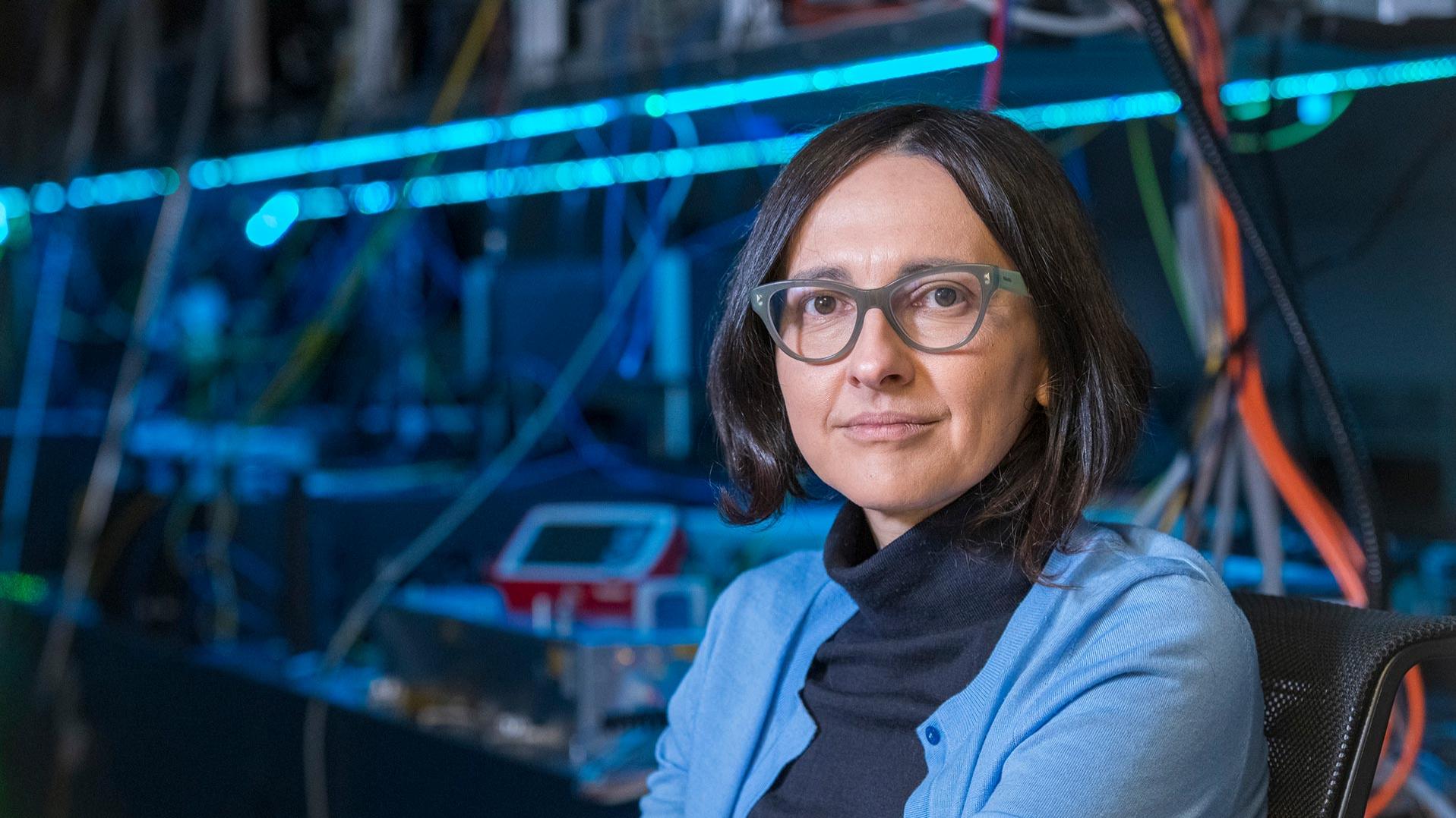 Professor Dr Jelena Vuckovic sits in a laboratory with technical equipment in the background. She is wearing a black turtleneck jumper and a light blue cardigan.