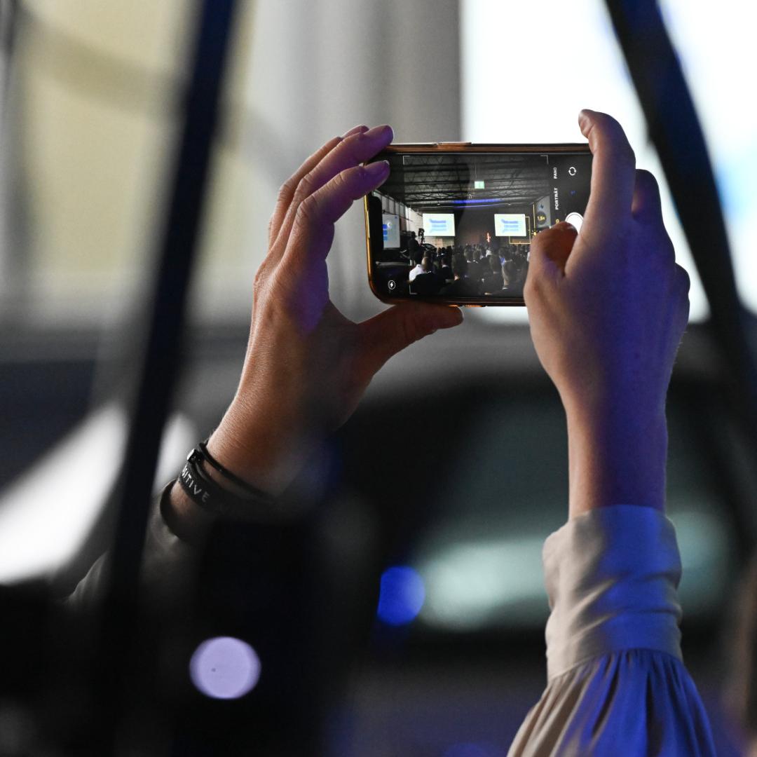 A smartphone is held up at an event in landscape orientation to capture a photograph of the events seen on stage