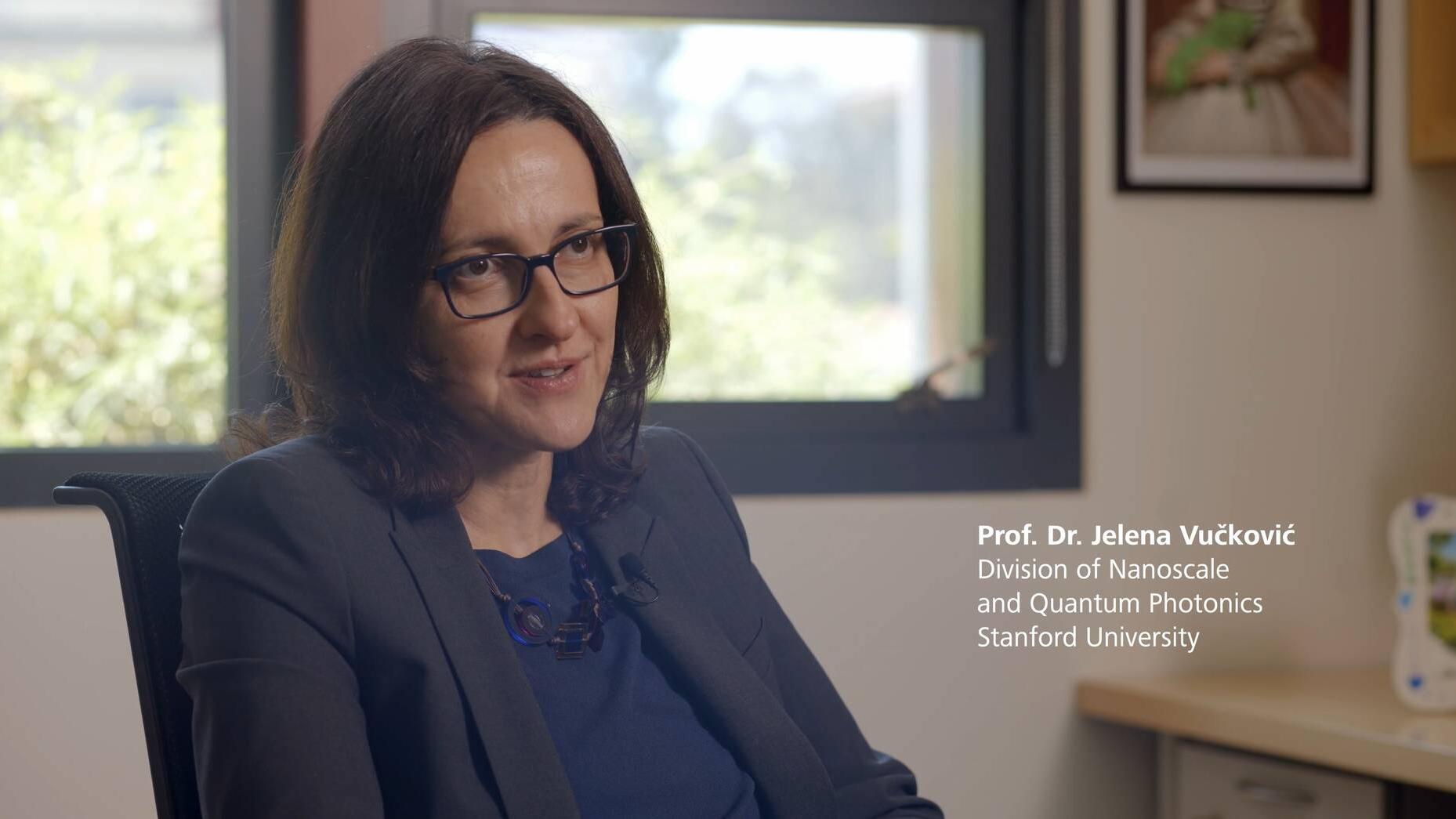 ZEISS Research Award Winner 2025: Prof. Dr. Jelena Vučković sitting in her office. She is wearing a dark blue blouse with a navy blue blazer. She has shoulder-length, dark brown hair and is wearing rectangular eyeglasses and is positioned at an angle to the camera.