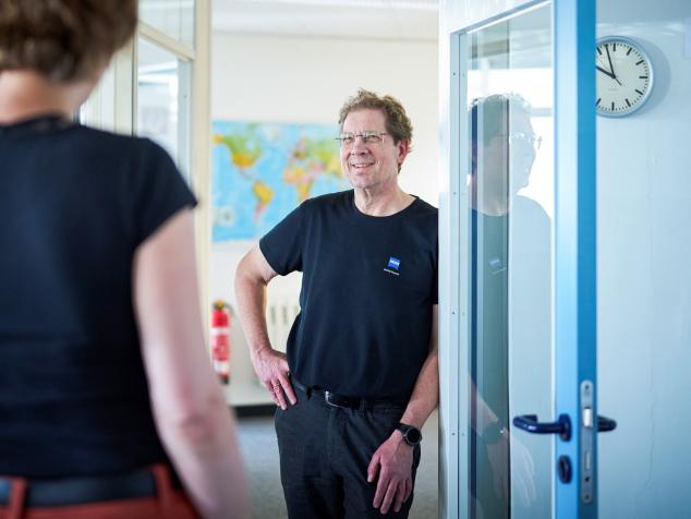 John Thomas Lakey stands in a doorway, smiling and talking to a woman with her back to the camera. A world map is visible behind him, and clocks showing different time zones are mounted on the wall to the right.