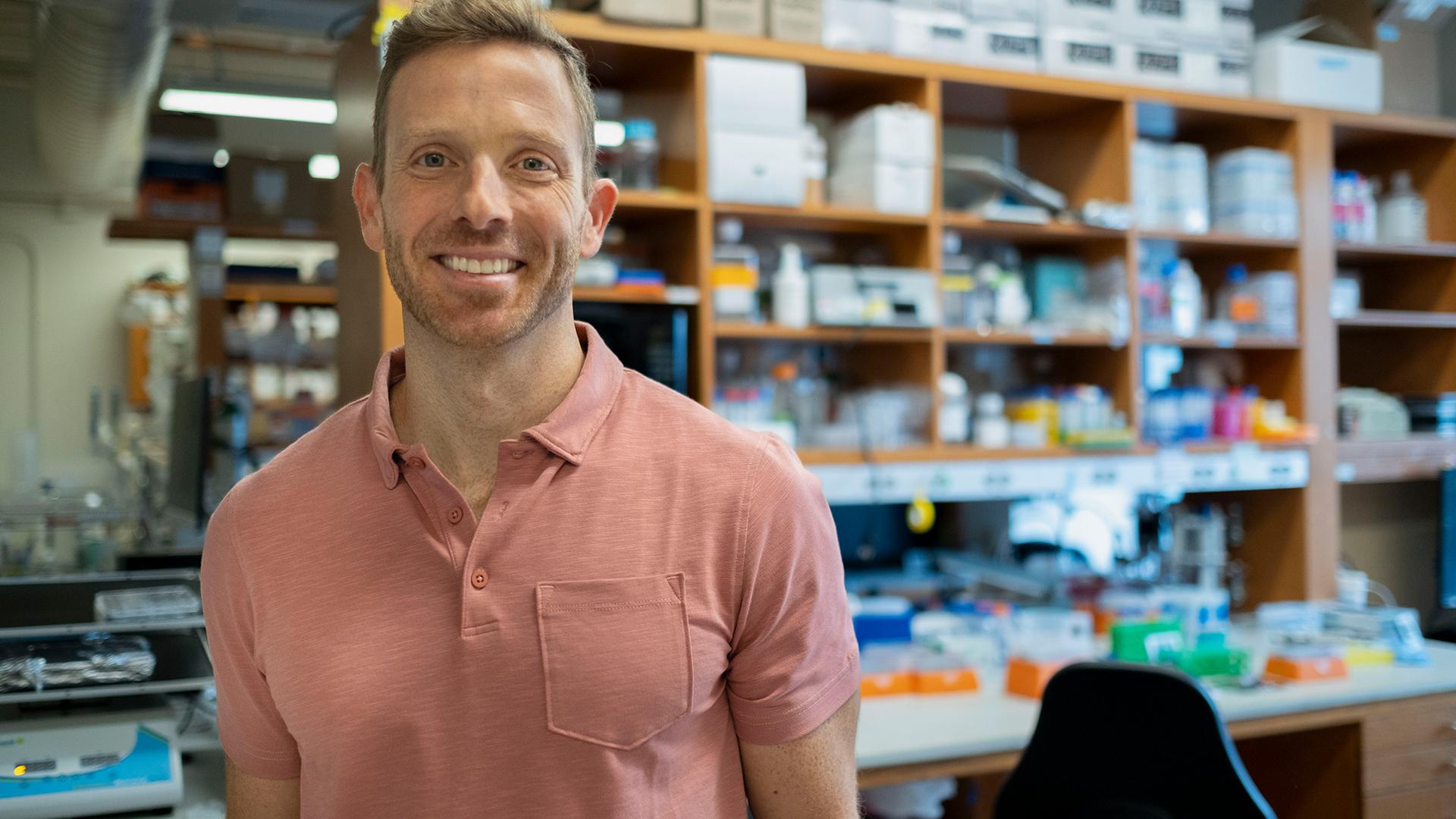 Prof. Benjamin L. Prosser stands in a laboratory filled with various scientific equipment and supplies on shelves behind him.