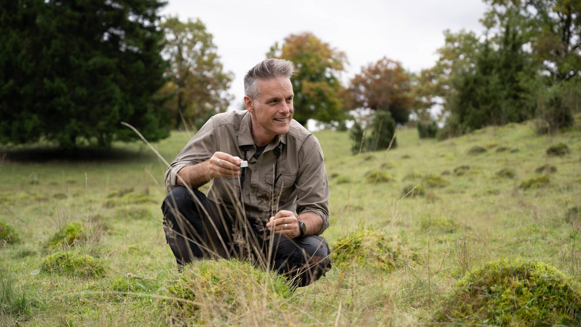 Nick Baker is perched in a meadow with lush green trees in the background. He is wearing a beige button-down-shirt and khaki working pants. He holds a small magnifying glass in his left hand and looks away from the camera at an angle.