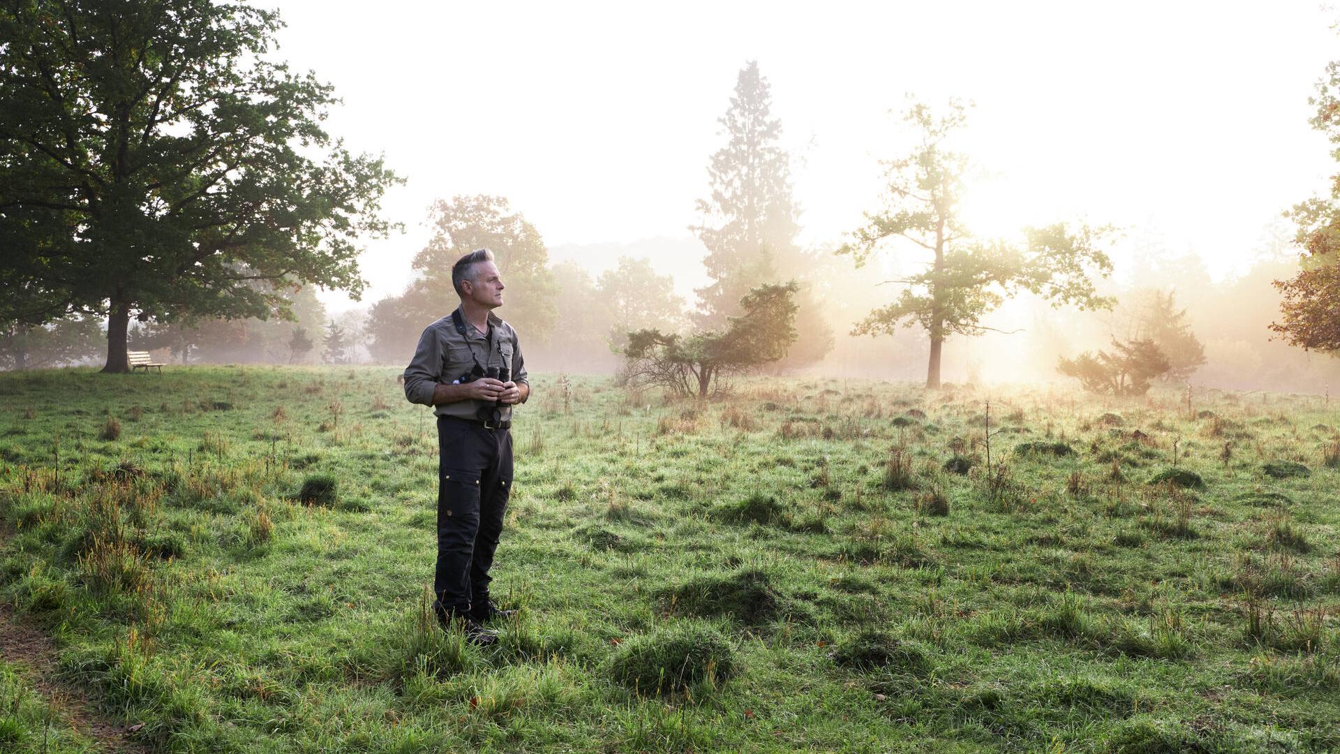 Nick Baker is standing in meadow. The surroundings are partially hidden by early morning fog which is illuminated by the rising sun.