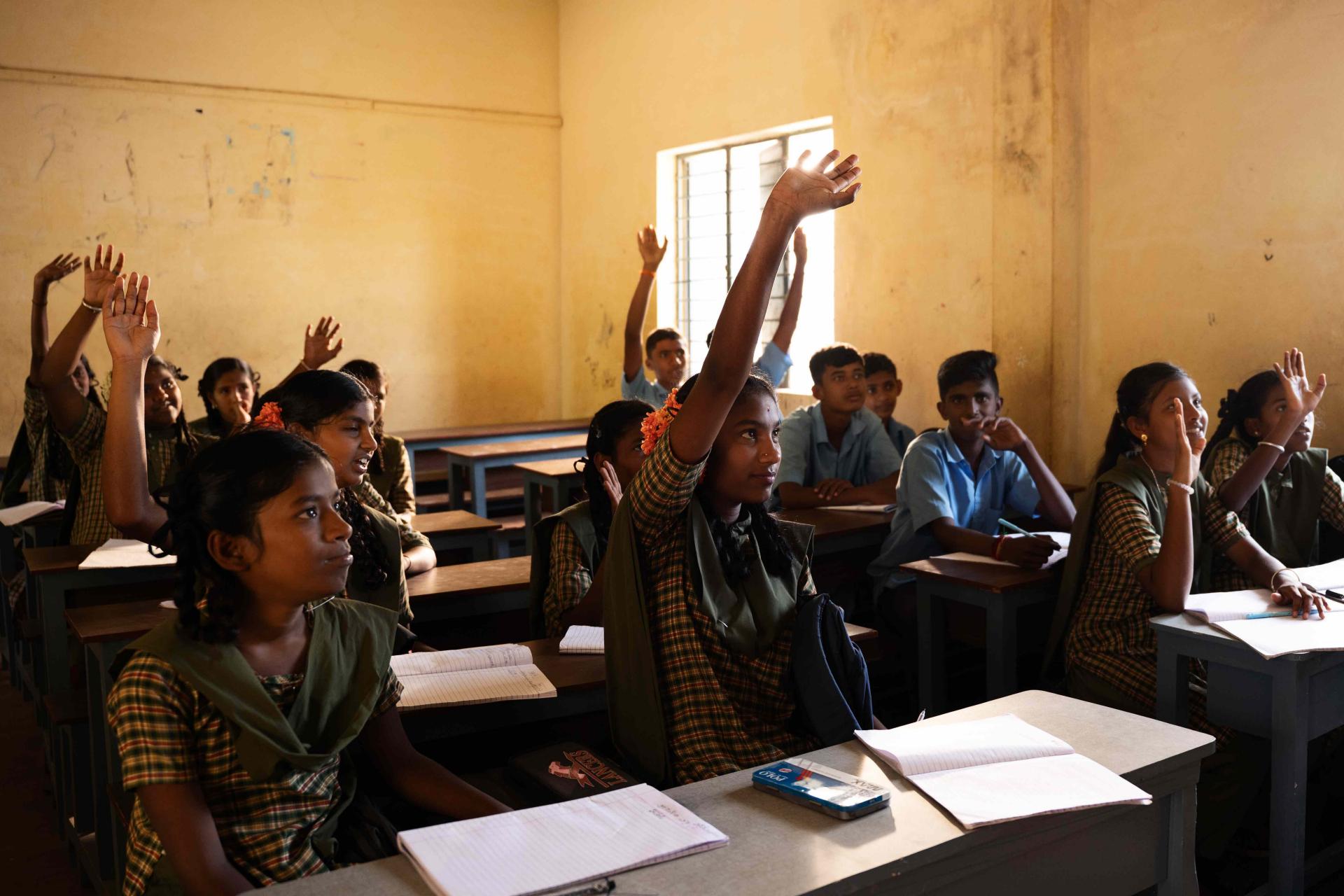 Children sitting on the benches at their desks. Some of them have raised their hands