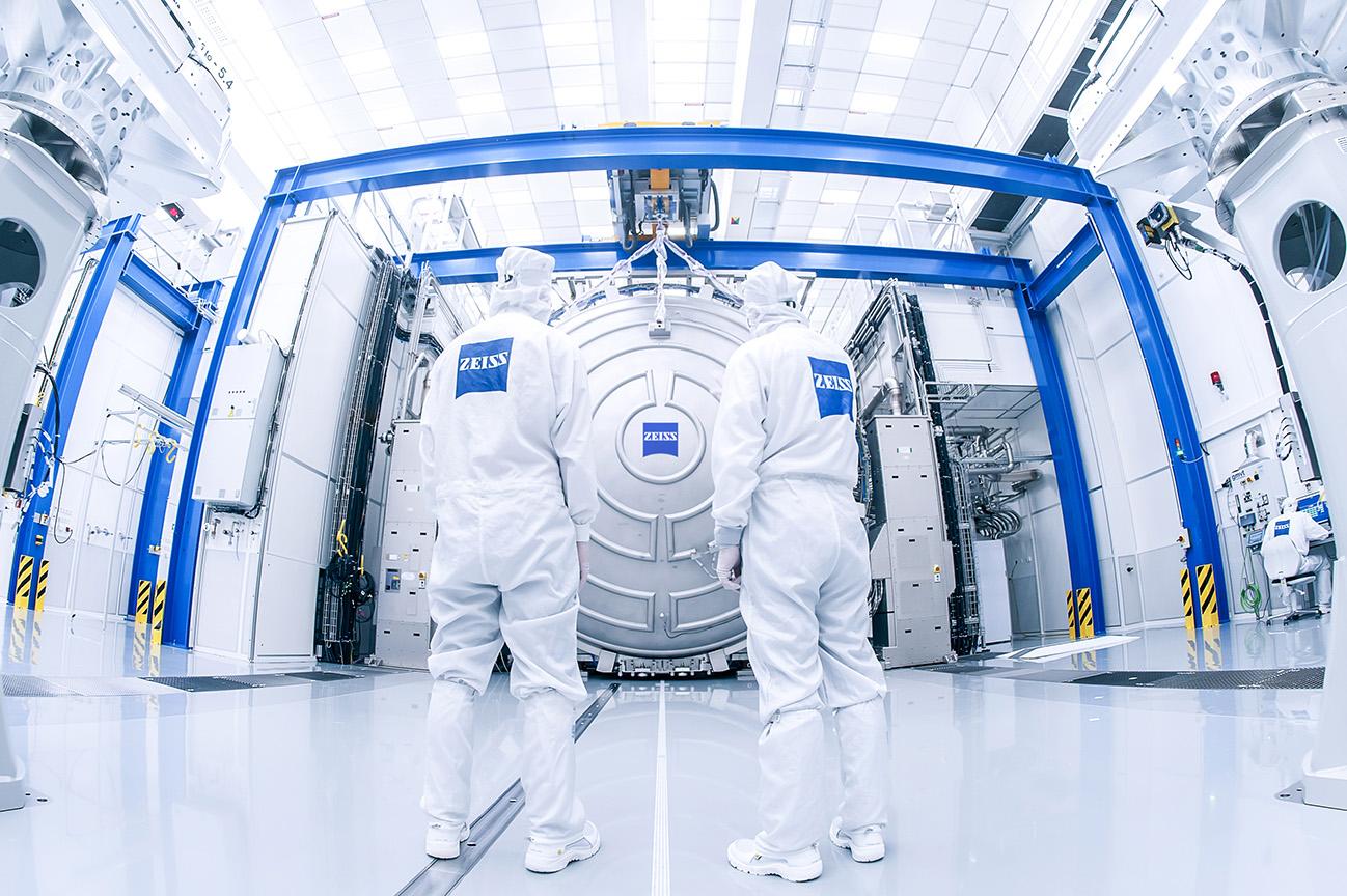 Two ZEISS employees in cleanroom clothing standing in front of a large vacuum chamber.