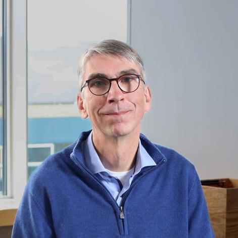 Portrait of Dr. Ulf Wilhelm. He is sitting on a stool in a community space in an office environment and looks straight at the camera