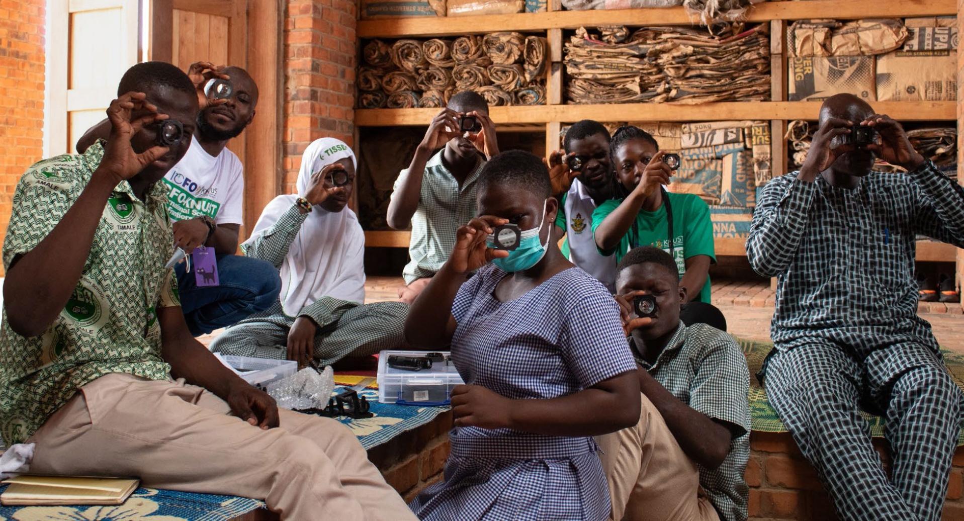Acht Schülerinnen und Schüler aus Ghana nehmen an einem Workshop teil, halten Linsen hoch und schauen hindurch.