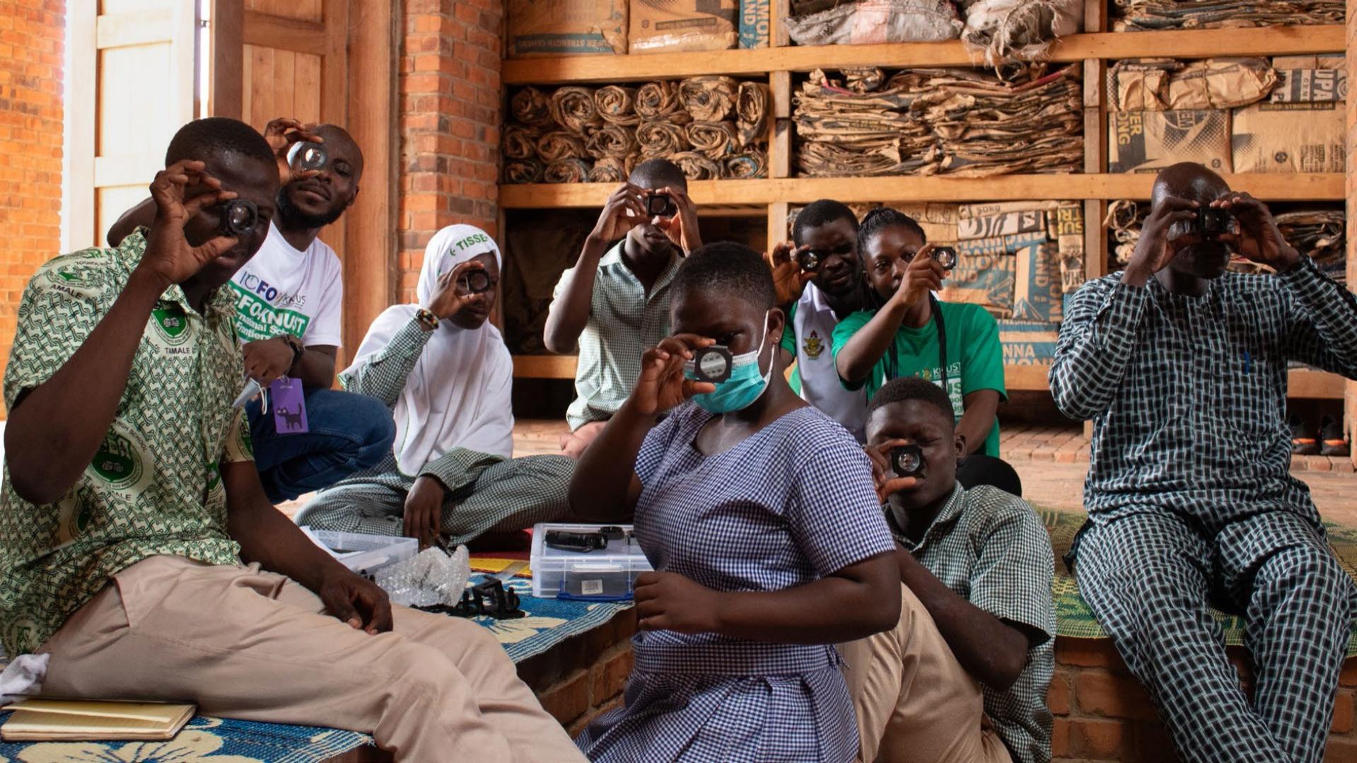 Acht Schülerinnen und Schüler aus Ghana nehmen an einem Workshop teil, halten Linsen hoch und schauen hindurch.