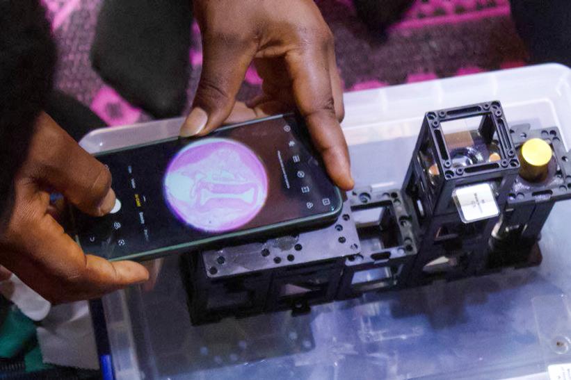 Nahaufnahme einer Hand, die ein Smartphone über ein modulares Mikroskop hält. Auf dem Smartphone-Bildschirm ist eine mikroskopische Ansicht sichtbar. Das Mikroskop befindet sich in einer transparenten Kunststoffbox.