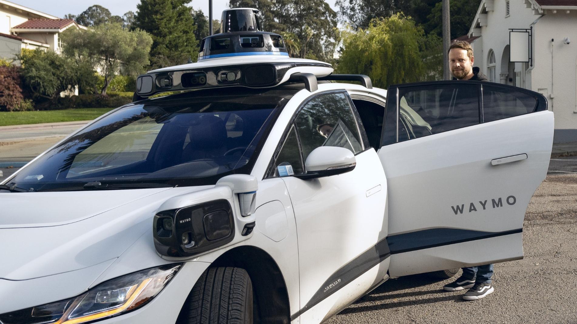 Ein autonomes Fahrzeug von Waymo steht auf einem Parkplatz, während ein Mann (Christoph Hoffmann) gerade die Beifahrertür öffnet. Die Technologie auf dem Fahrzeugdach und an den Seiten, wie Sensoren und Kameras, ist gut sichtbar.