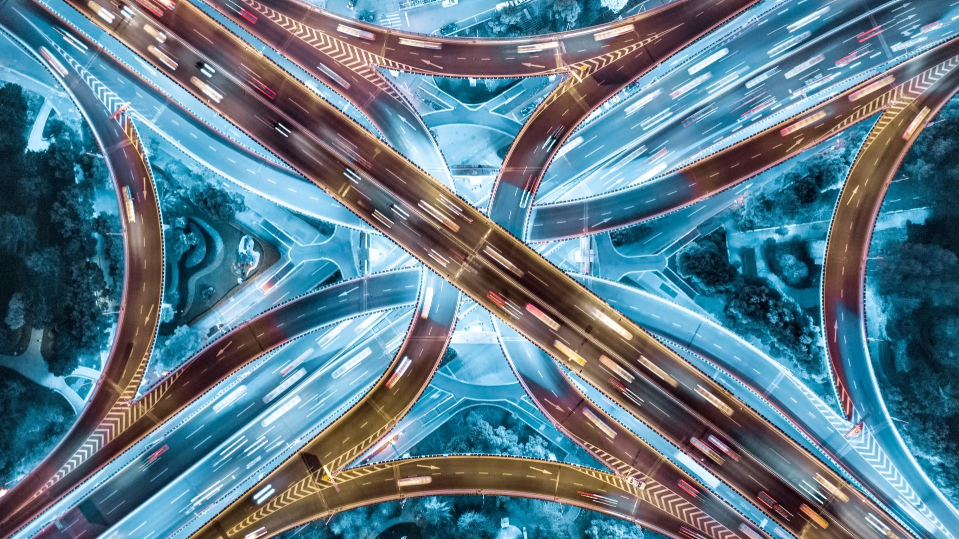 Luftaufnahme eines komplexen Autobahnkreuzes bei Nacht, beleuchtet durch Scheinwerfer der Fahrzeuge, die als Lichtstreifen sichtbar sind. Die Überführungen und Kurven bilden ein symmetrisches Muster, umgeben von Bäumen und kleinen Grünflächen.