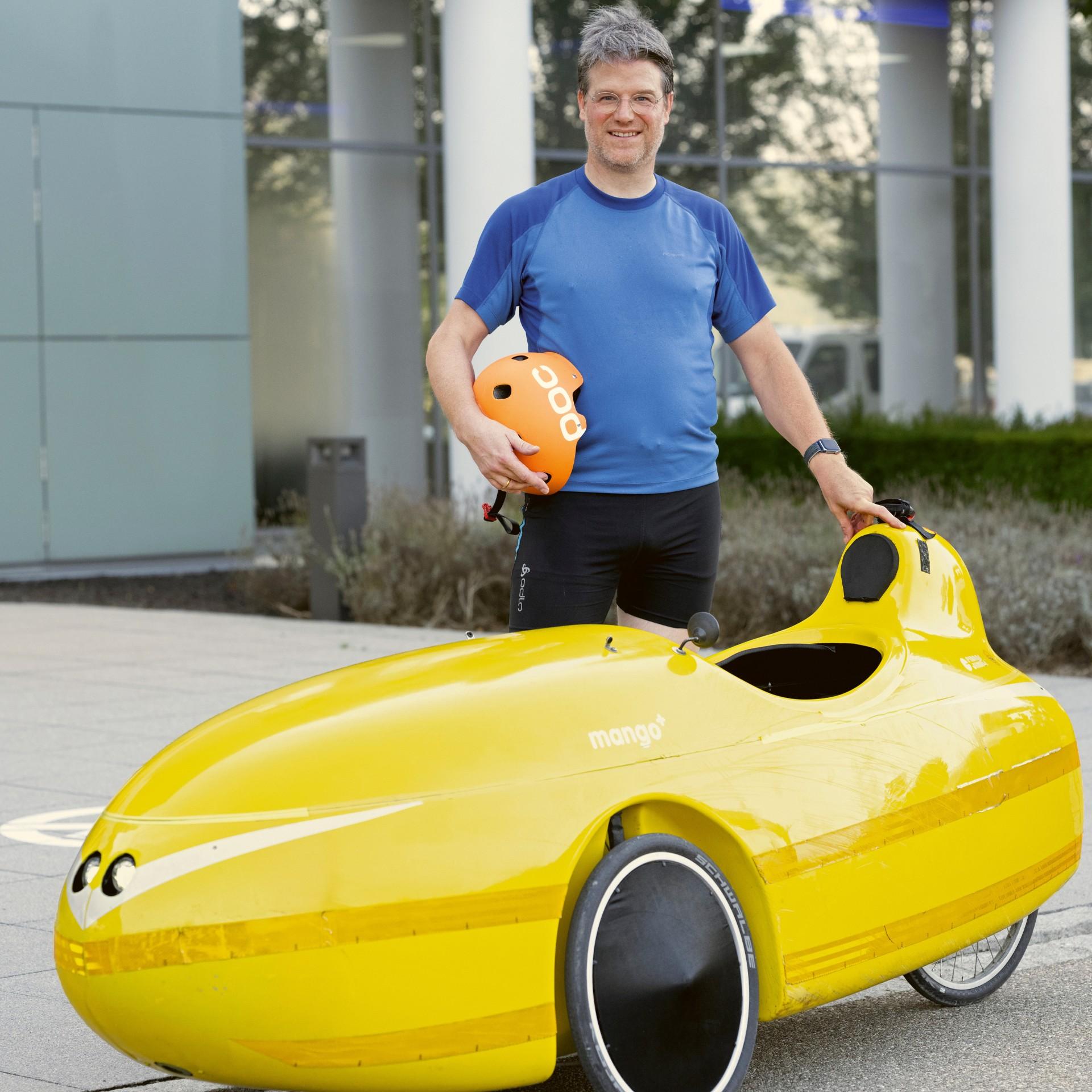 Dr. Bertram Klein in sportlicher Kleidung steht neben einem gelben Velomobil des Modells „Mango“, das für nachhaltige Fortbewegung entwickelt wurde. Er hält einen orangefarbenen Fahrradhelm in der Hand, lächelt und blickt in die Kamera.