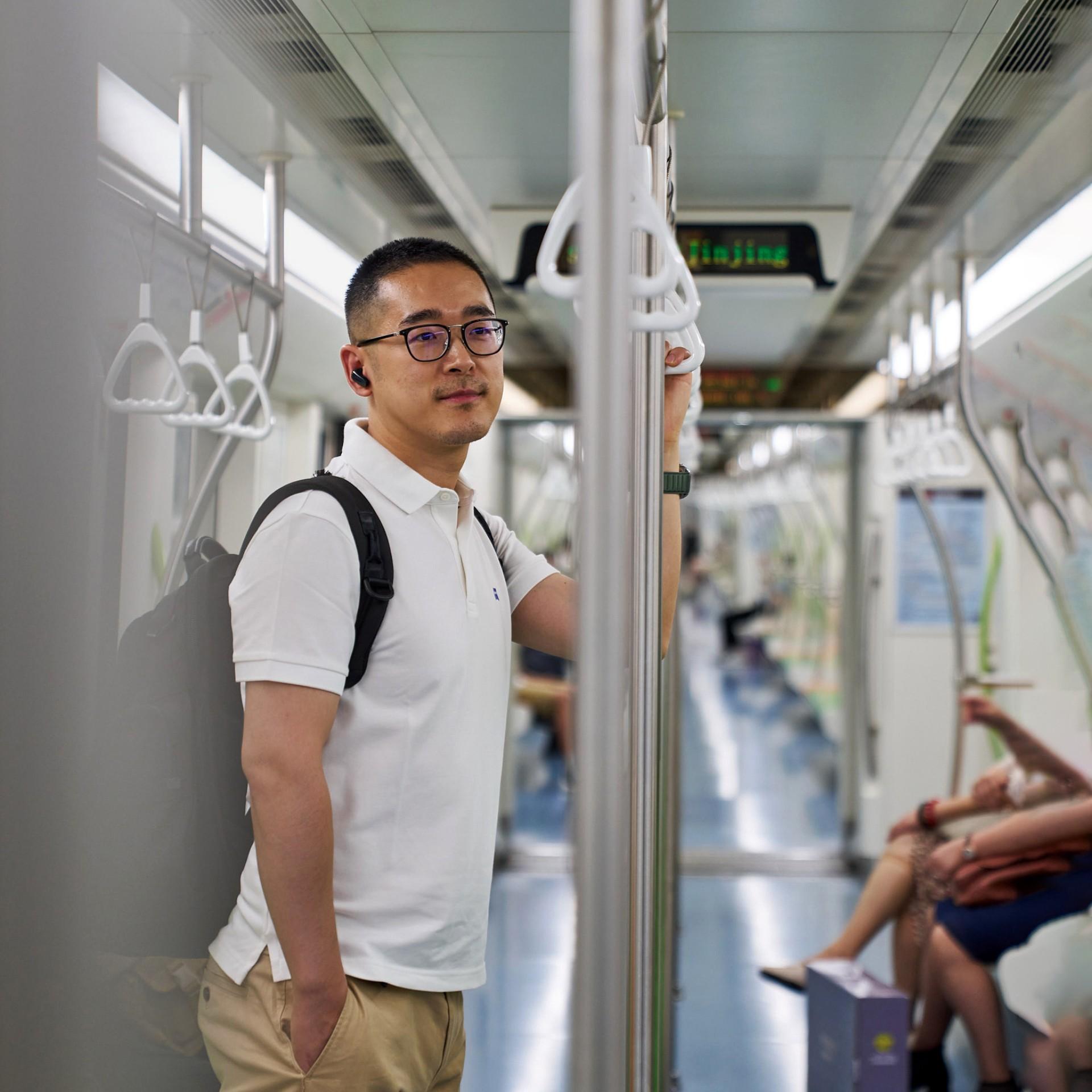 Victor Miao in einem weißen Polohemd mit Rucksack steht in einer U-Bahn, hält sich an einer Stange fest und blickt in die Kamera. Im Hintergrund sitzen weitere Passagiere, und die Inneneinrichtung der U-Bahn ist sichtbar.