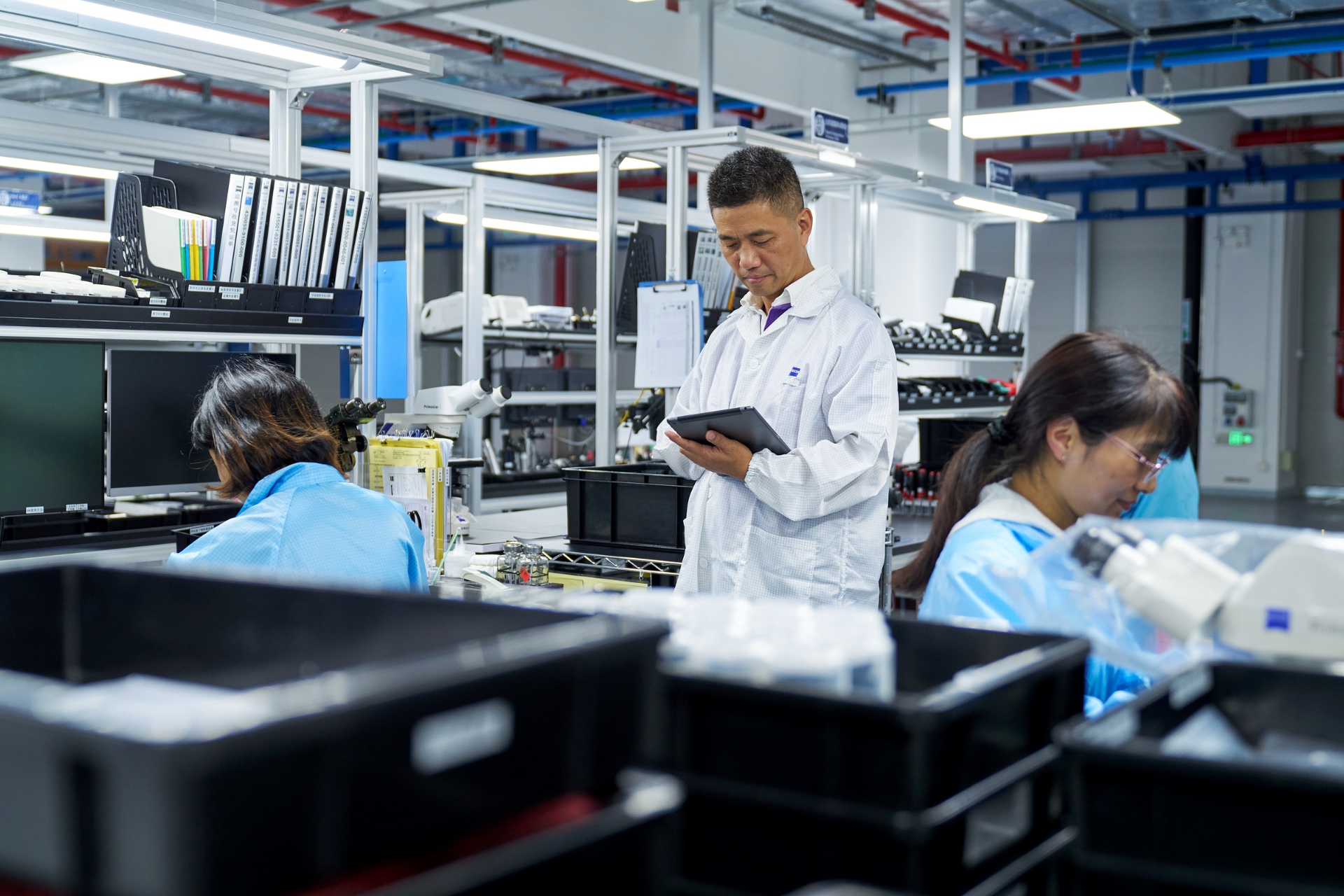 Produktionsingenieur Ke Yong steht mit einem Tablet in einem Produktionsbereich, während zwei Mitarbeiterinnen im Hintergrund ihren Tätigkeiten nachgehen.