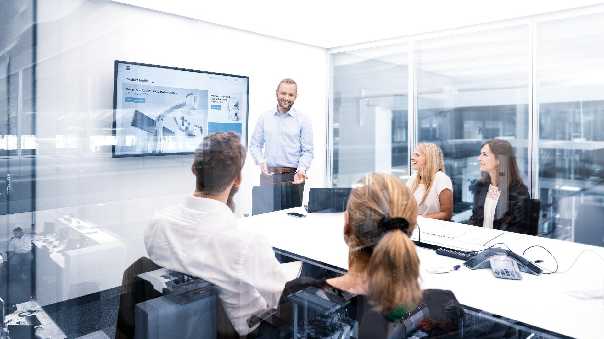 A man in a shirt gives a presentation in a modern conference room. He is standing in front of four people, who are sitting at a desk and attentively listening to him. In the background are large glass partitions providing a view of the rest of the office. A presentation with the title 'Product highlights' can be seen on a screen on the wall.