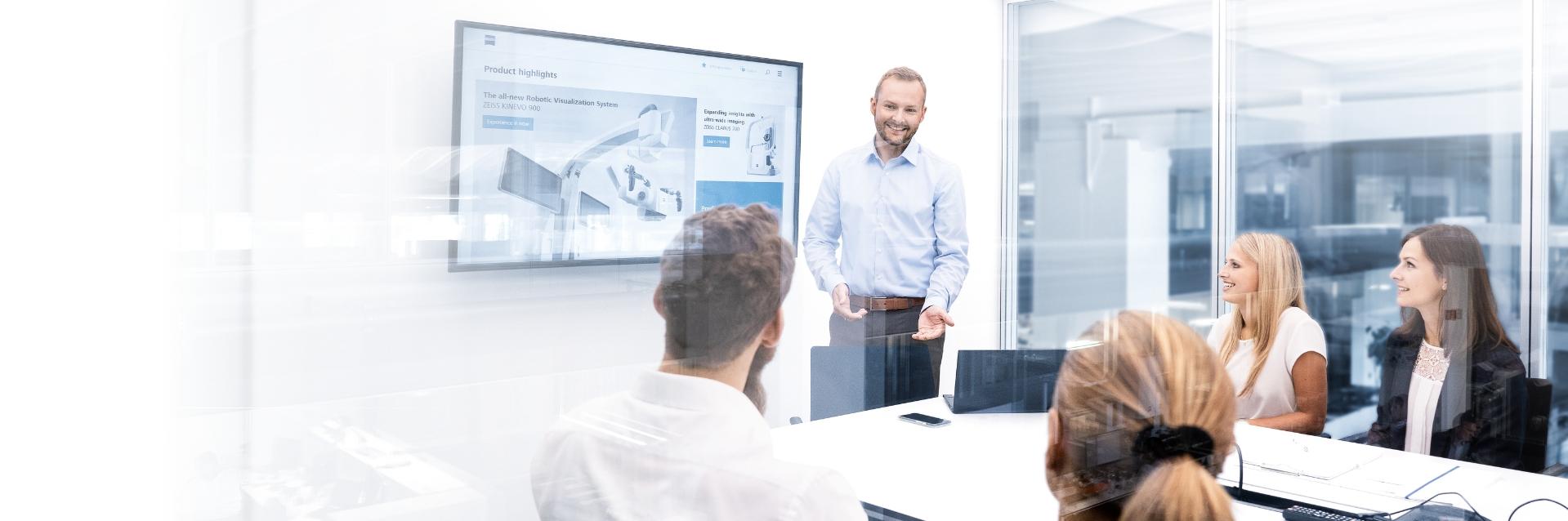 A man in a shirt gives a presentation in a modern conference room. He is standing in front of four people, who are sitting at a desk and attentively listening to him. In the background are large glass partitions providing a view of the rest of the office. A presentation with the title 'Product highlights' can be seen on a screen on the wall.