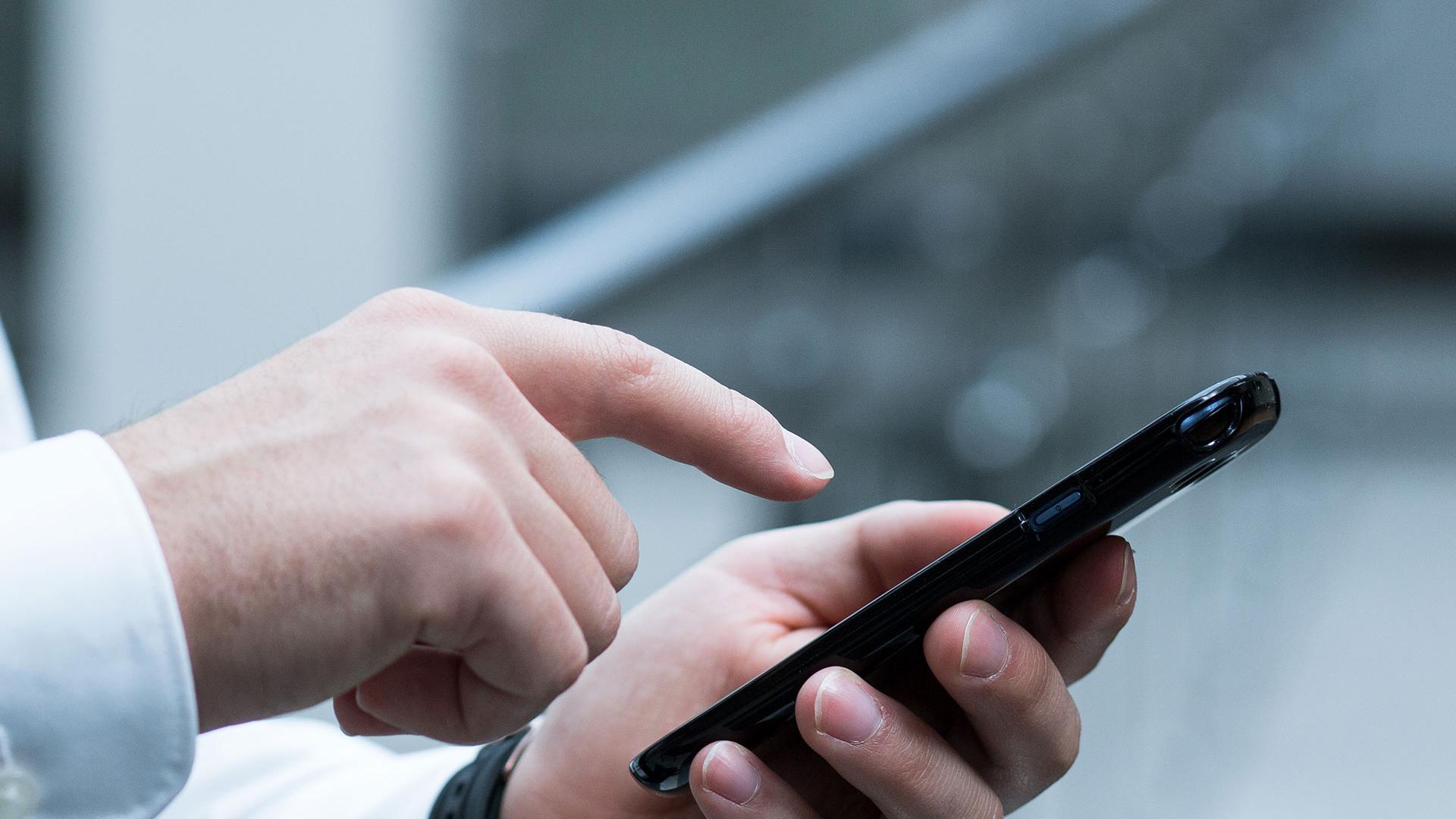 A close-up shot of a person's hand holding a smartphone, while they point at the screen with their other hand.