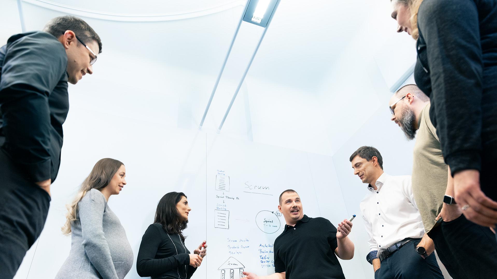 Eine Gruppe von Personen steht vor einem Whiteboard. Ein Mann hält eine Präsentation, während die anderen zuhören.