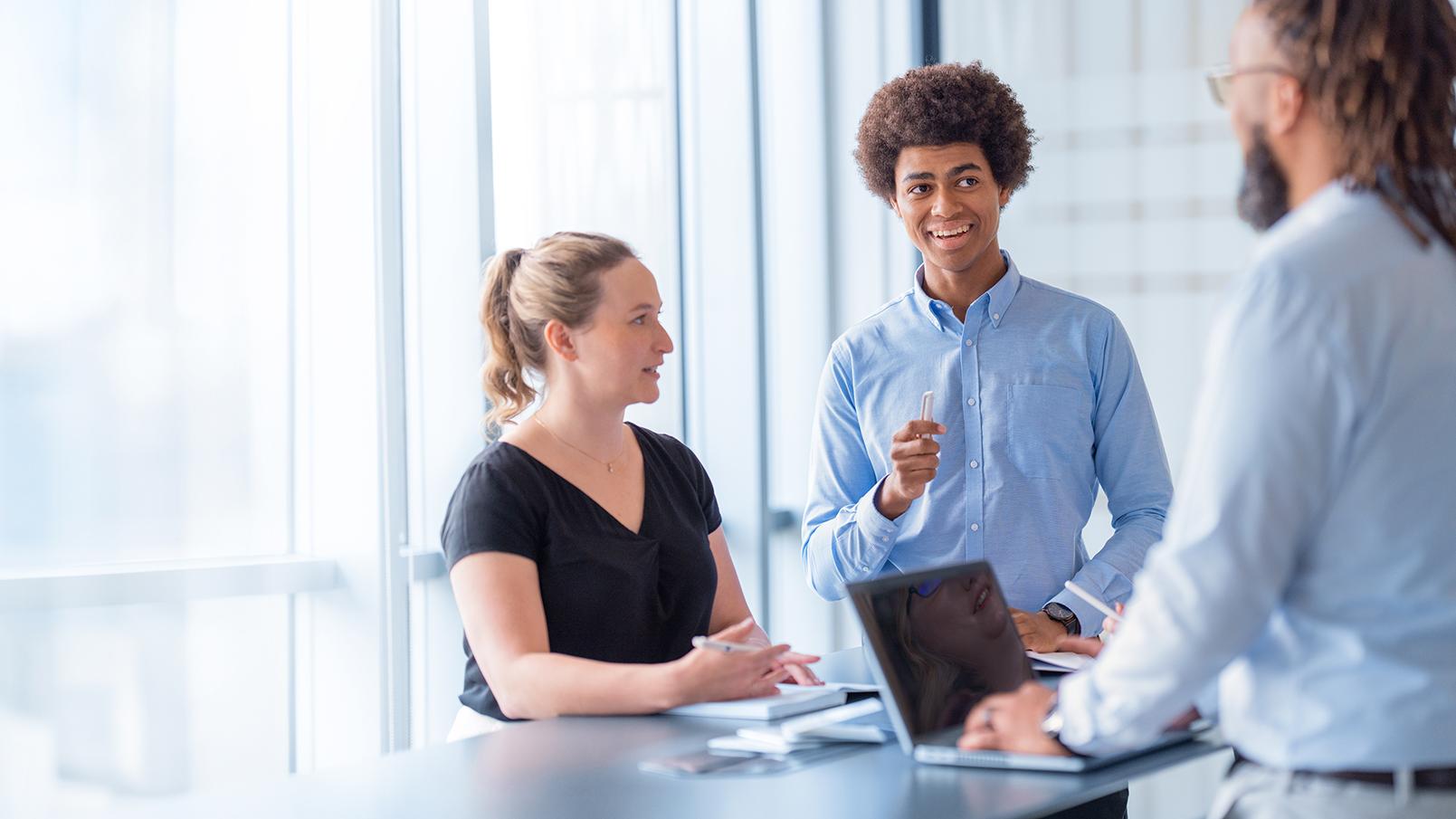 Three people stand at a desk in a modern office and chat.