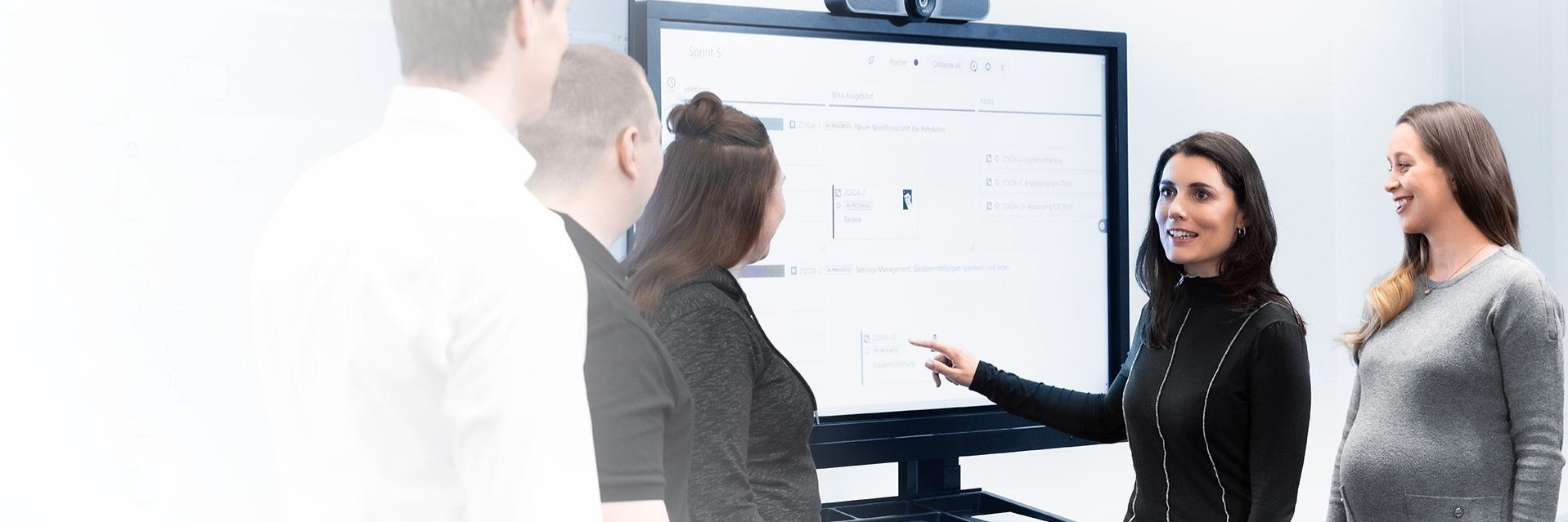 A group of five people stand in front of two large screens in a modern conference room. A woman is giving a presentation and the audience listens and looks at the screens featuring diagrams.
