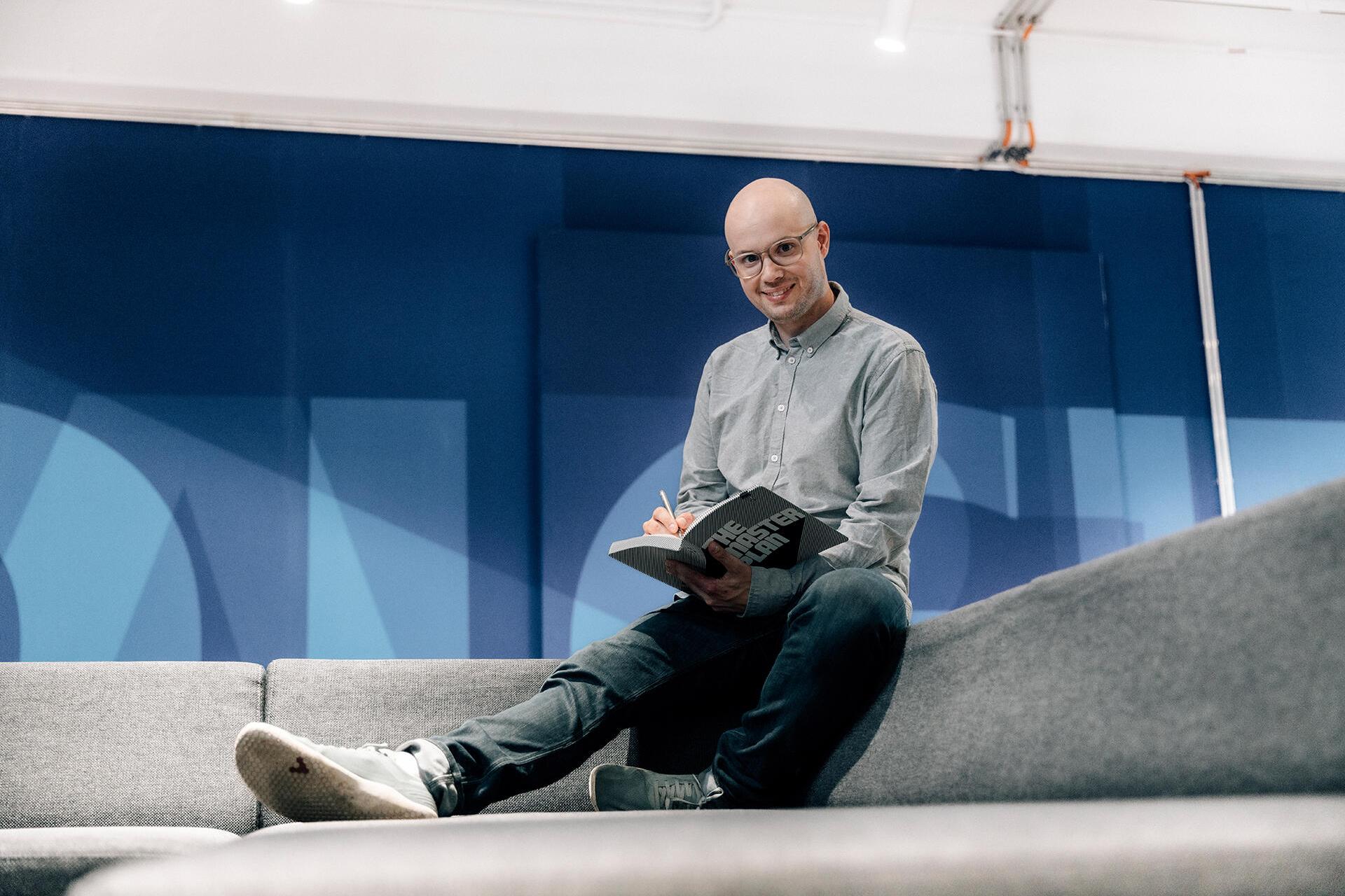 Emanuel sits, writes in a book and looks at the camera.