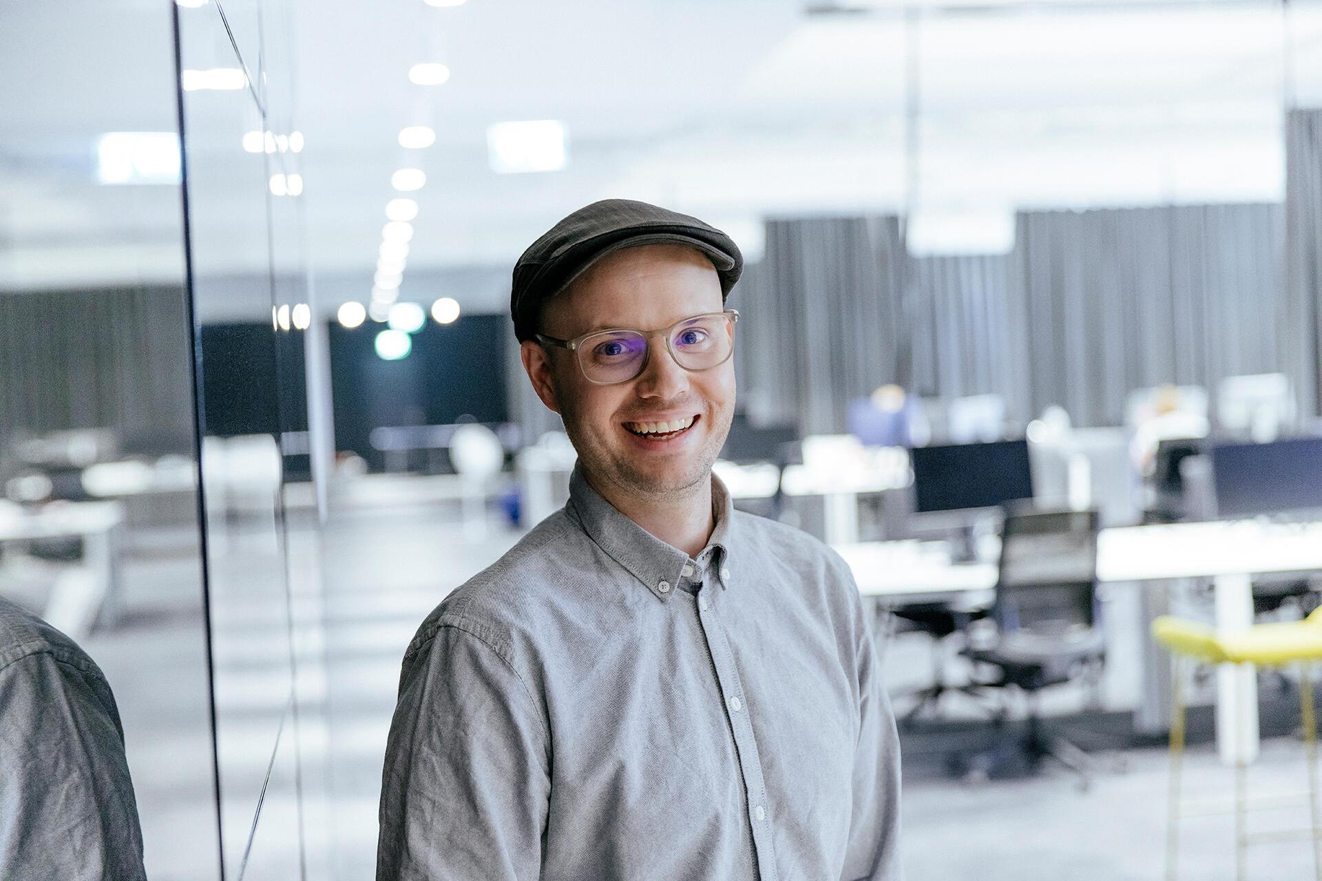 Emanuel stands in a modern office. He smiles and looks at the camera. 