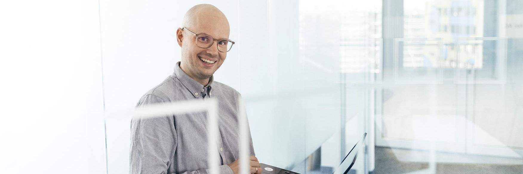 Emanuel stands in a modern office with glass partitions. He holds a laptop in his hand, looks at the camera and smiles.
