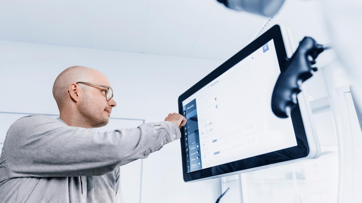 Emanuel steht an einem medizinischen ZEISS-Gerät im Operationssaal. Er konzentriert sich auf den Bildschirm, während er den Touchscreen des Geräts bedient.
