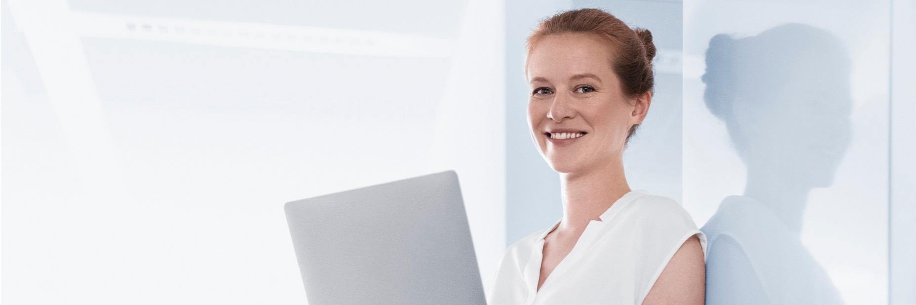 Franziska steht in einem hellen Raum, sie hält einen Laptop in der Hand und schaut in die Kamera.
