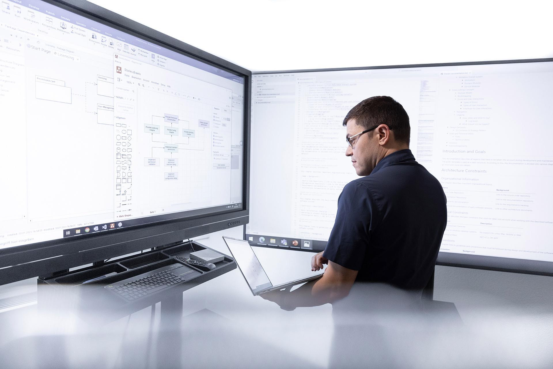 Hendrik works on a laptop while standing in front of two large monitors. The screens display a mixture of flowcharts and programming code.