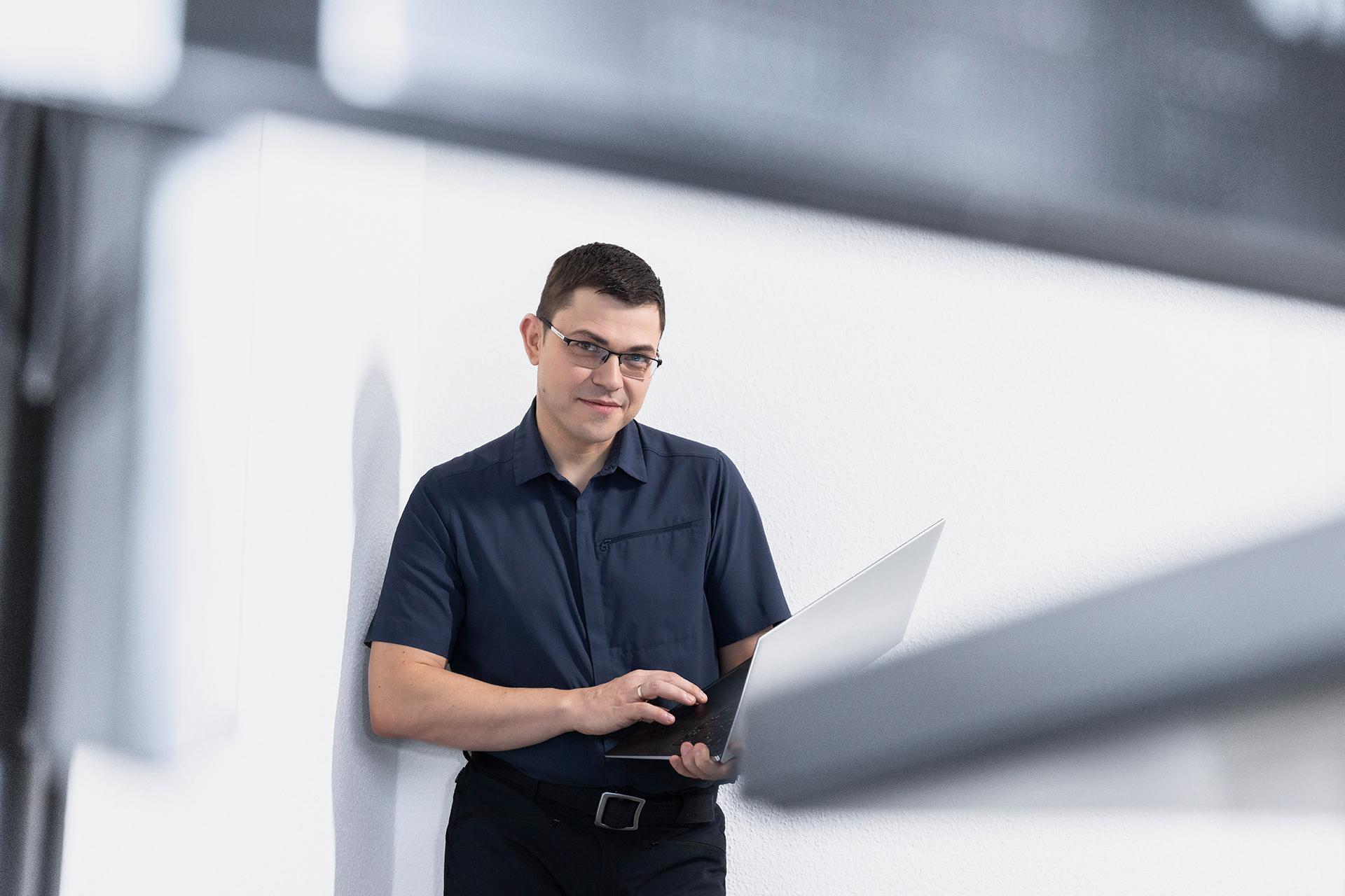 Hendrik leans against a white wall while holding an open laptop in his hands.