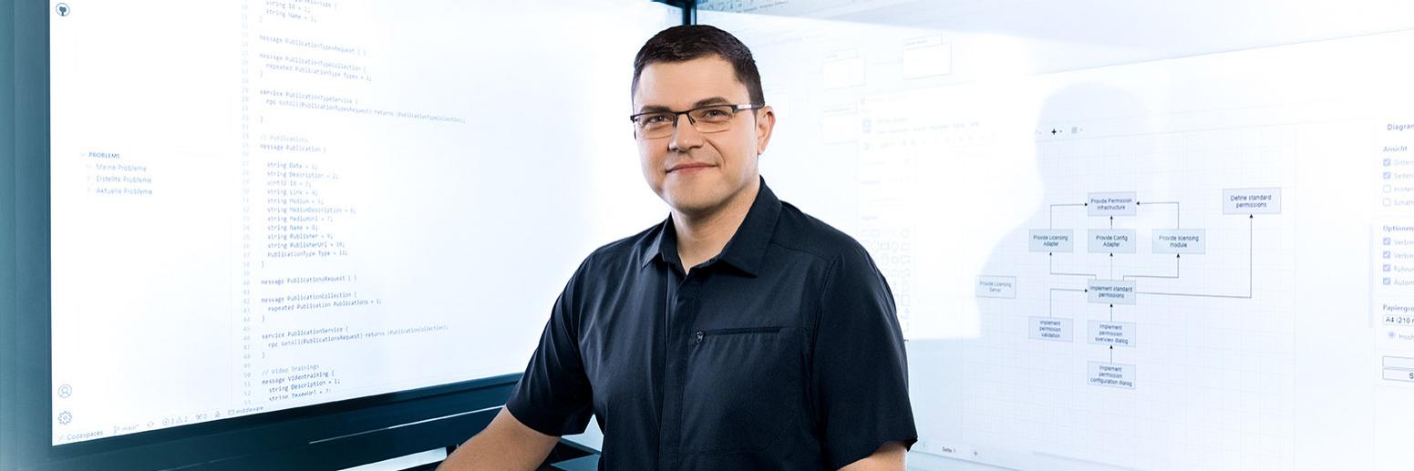 Hendrik stands in front of two large screens displaying technical diagrams and code.