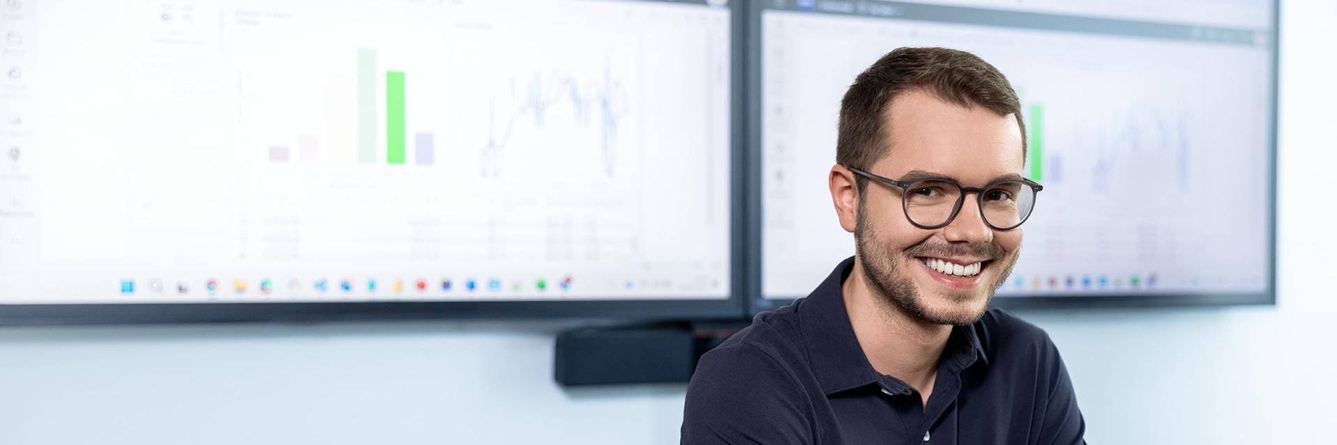 Julian looks relaxed and laughs as he sits in front of two monitors displaying a dashboard with various analytical data.