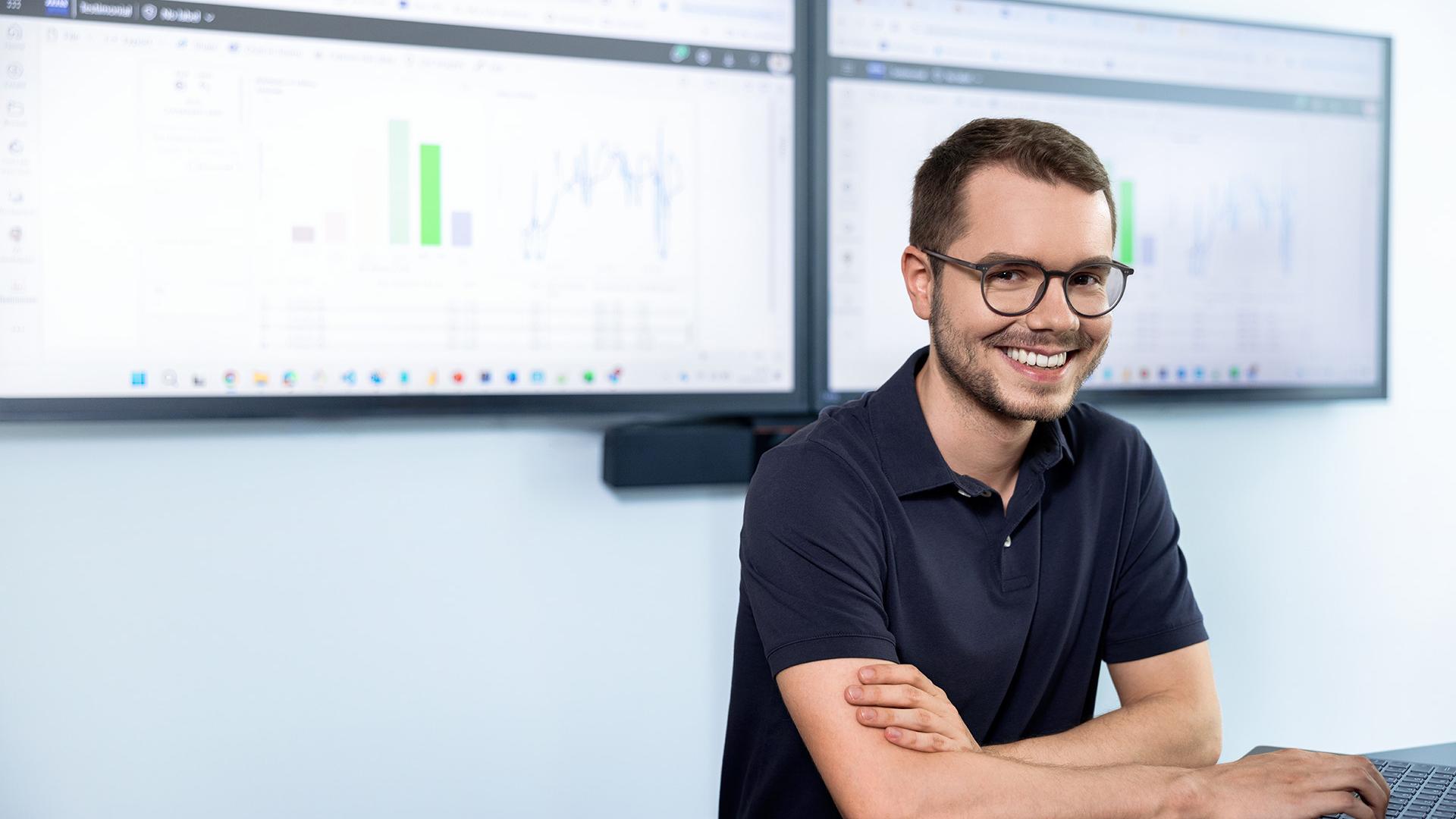Julian looks relaxed and laughs as he sits in front of two monitors displaying a dashboard with various analytical data.