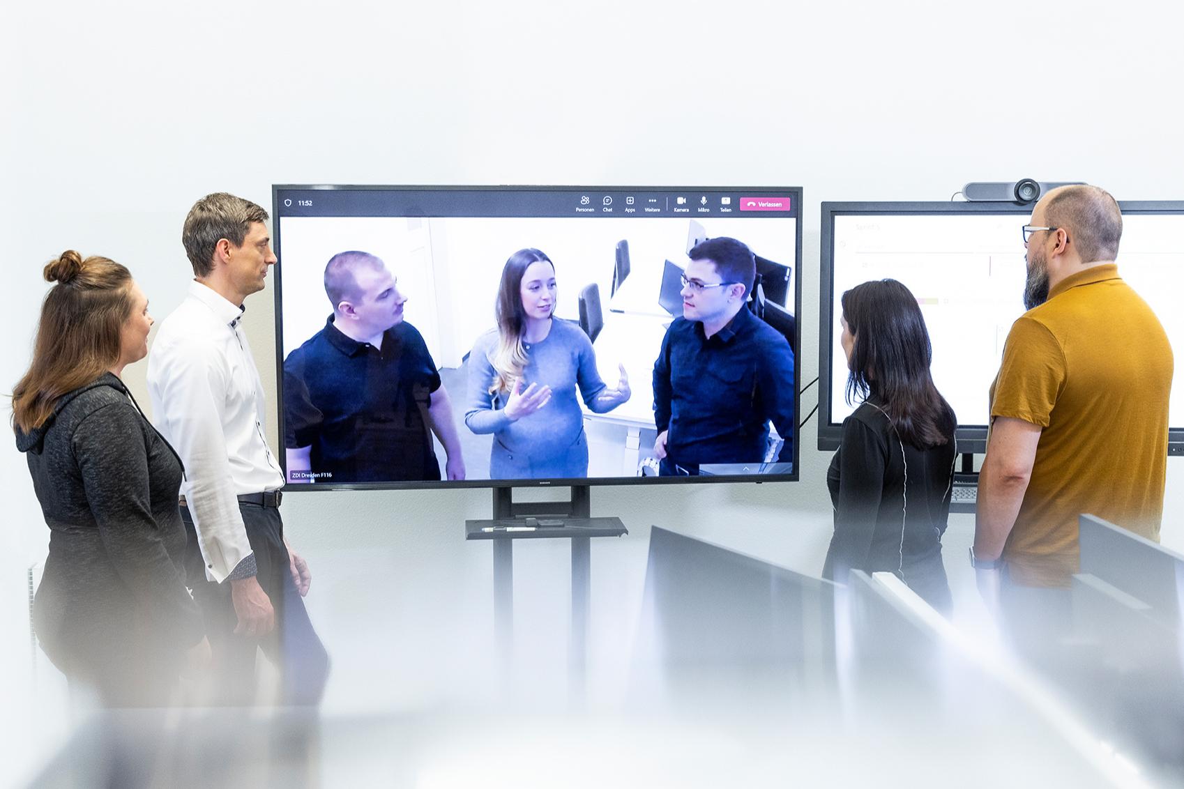 A team stands in front of a digital whiteboard and has an online meeting with Karolin and two other colleagues