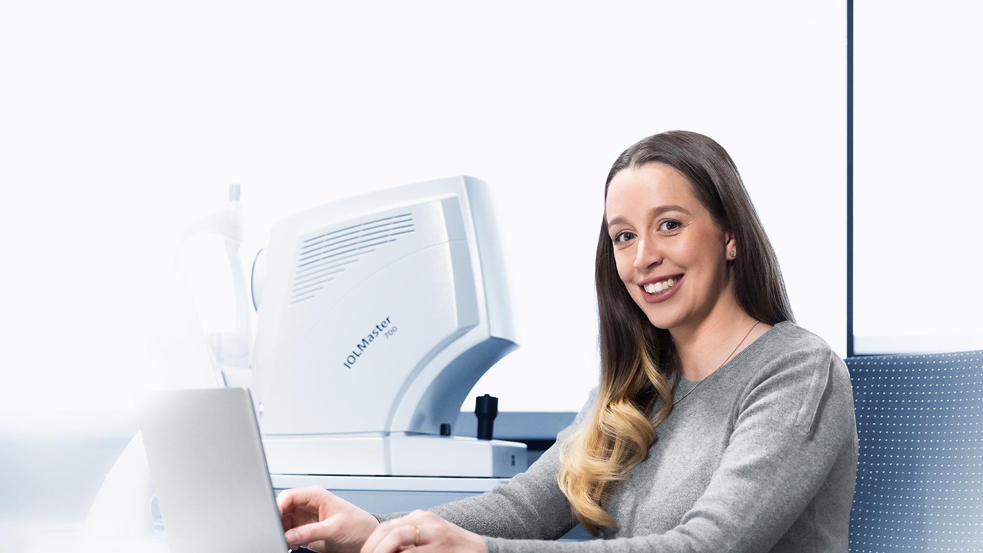 Karolin works on a laptop in a bright office. She looks warmly at the camera. The ZEISS IOLMaster 700 can be seen in the background