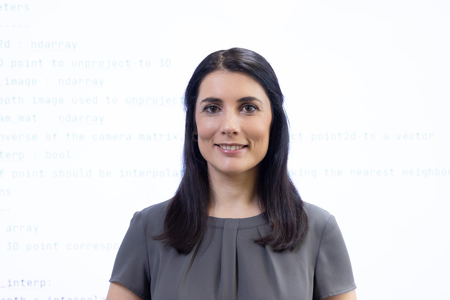 Sabrina stands in front of a large screen and smiles at the camera. Code is displayed on the screen in the background