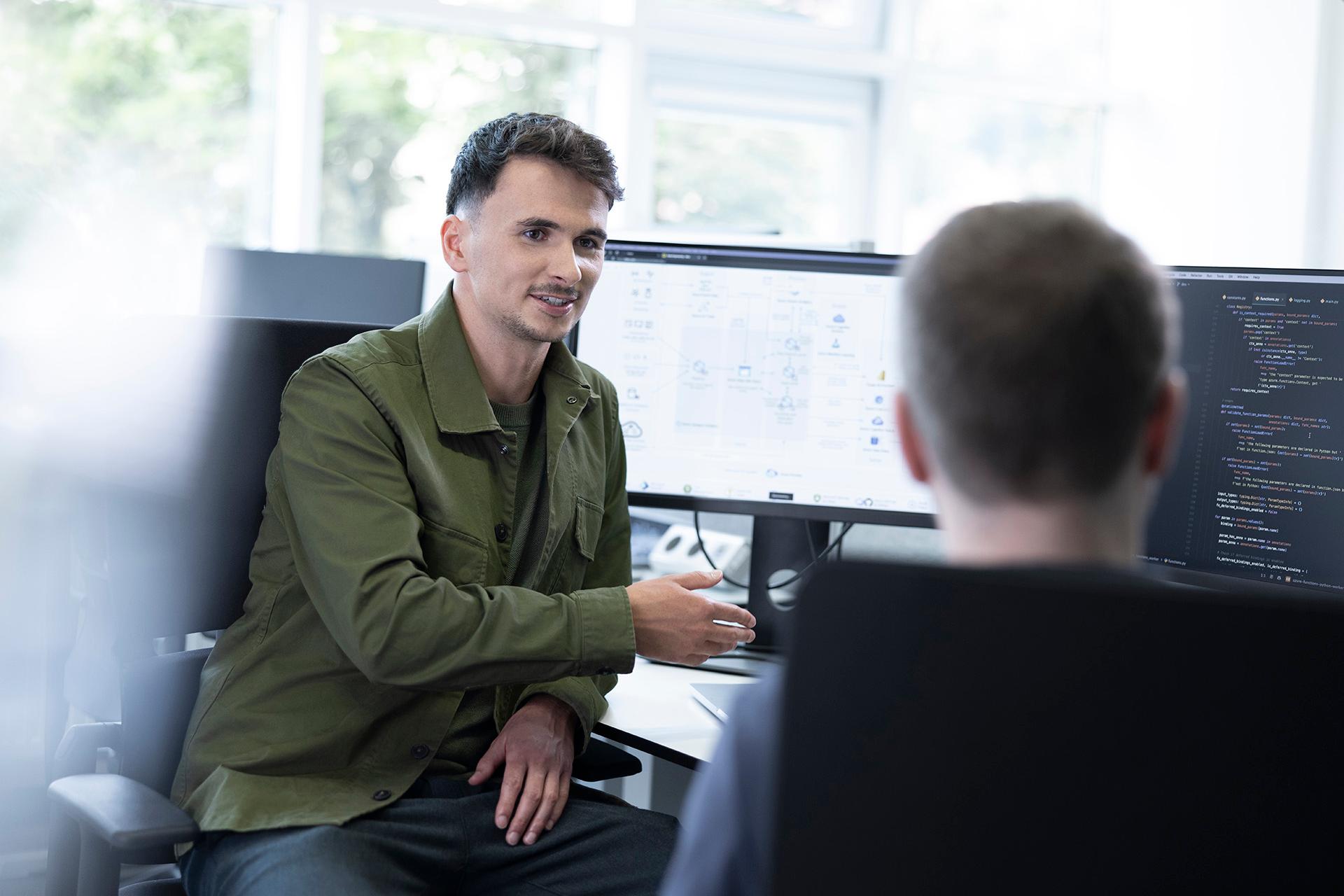 Simon is sitting in front of his computer at his desk and chatting to a male colleague.