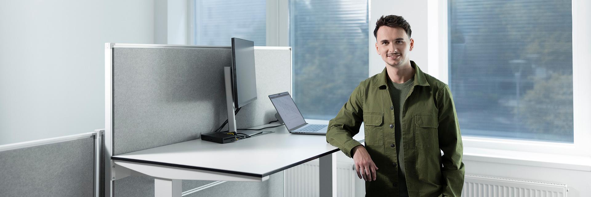Simon is standing at his desk, smiling and leaning in a relaxed stance.