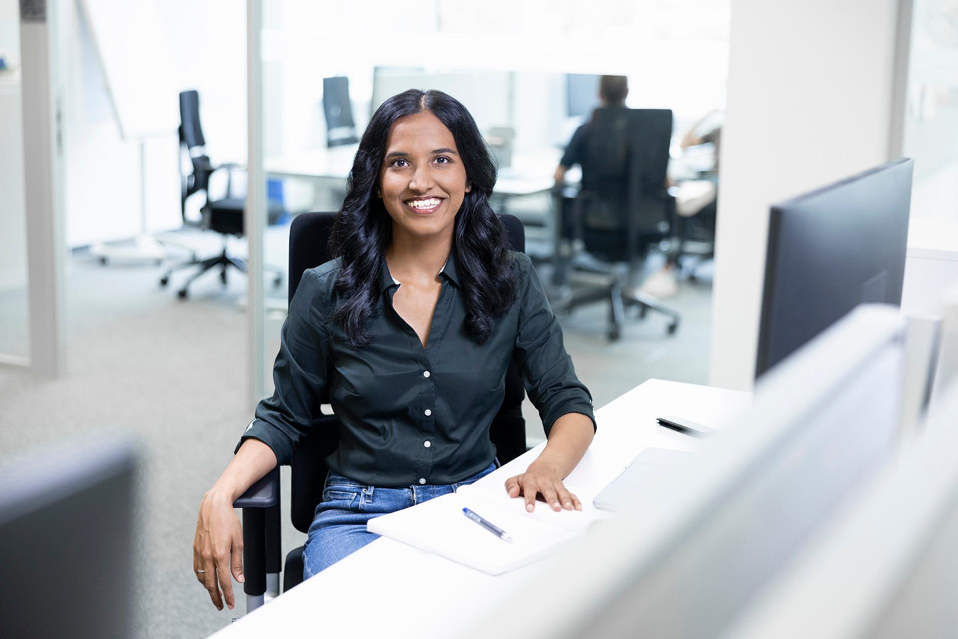 Varsha is sitting and smiling at her desk in a bright, spacious, glass-walled office.