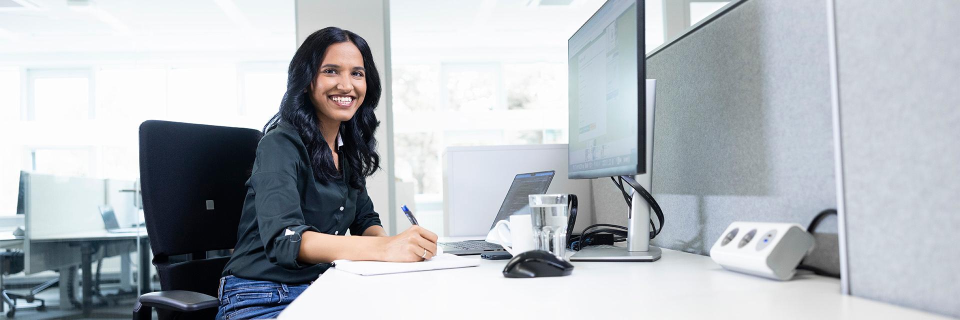 Varsha is sitting and smiling at her desk in her glass-walled office.