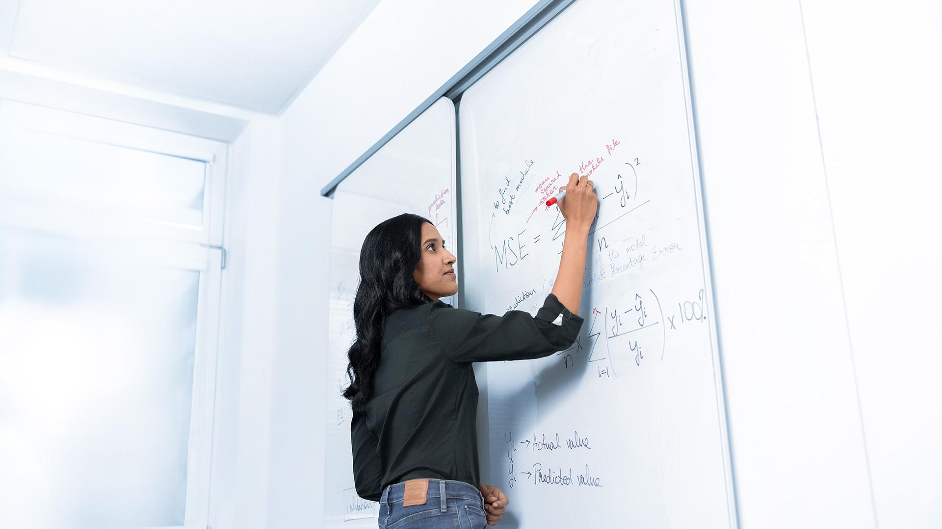 Varsha is standing in front of a whiteboard and scribbling math notes for data analysis.