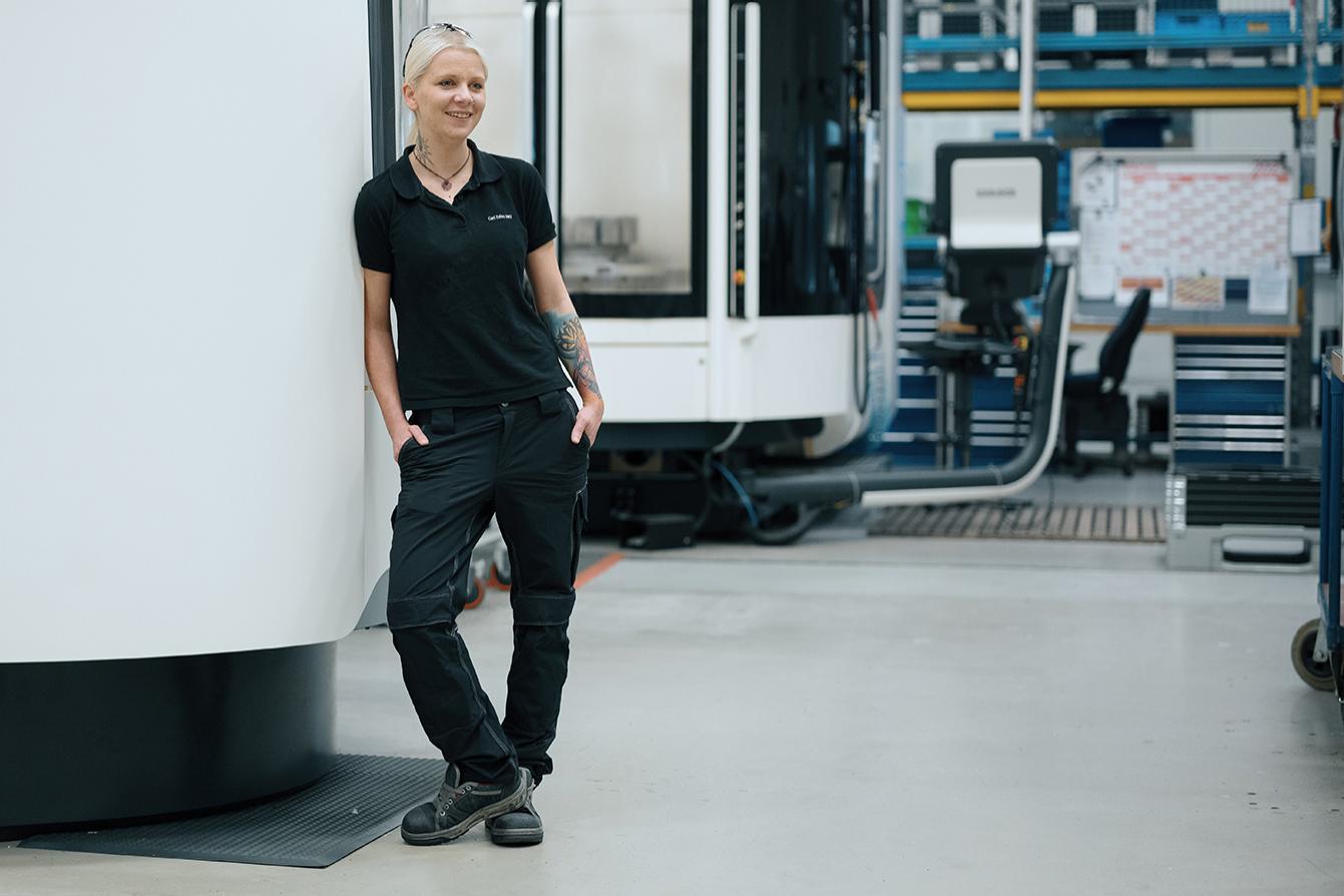 Melanie leans on a large white machine and smiles. She has black workwear on. Other machines can be seen in the background.