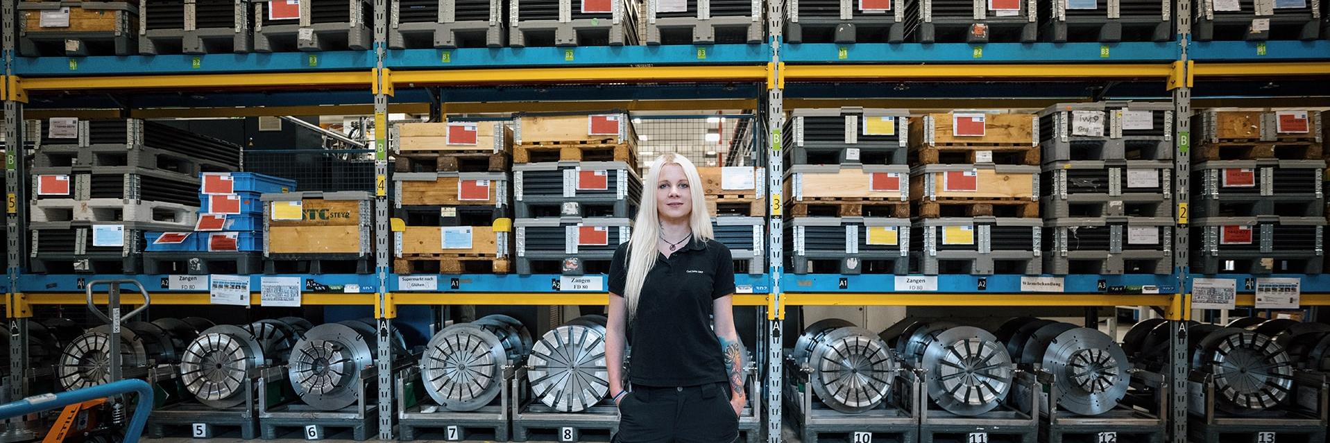 Melanie stands in a warehouse and looks at the camera. She has black workwear on. Various metal shelves with pallets on them can be seen in the background. Large, round metal parts are stored at the very bottom.