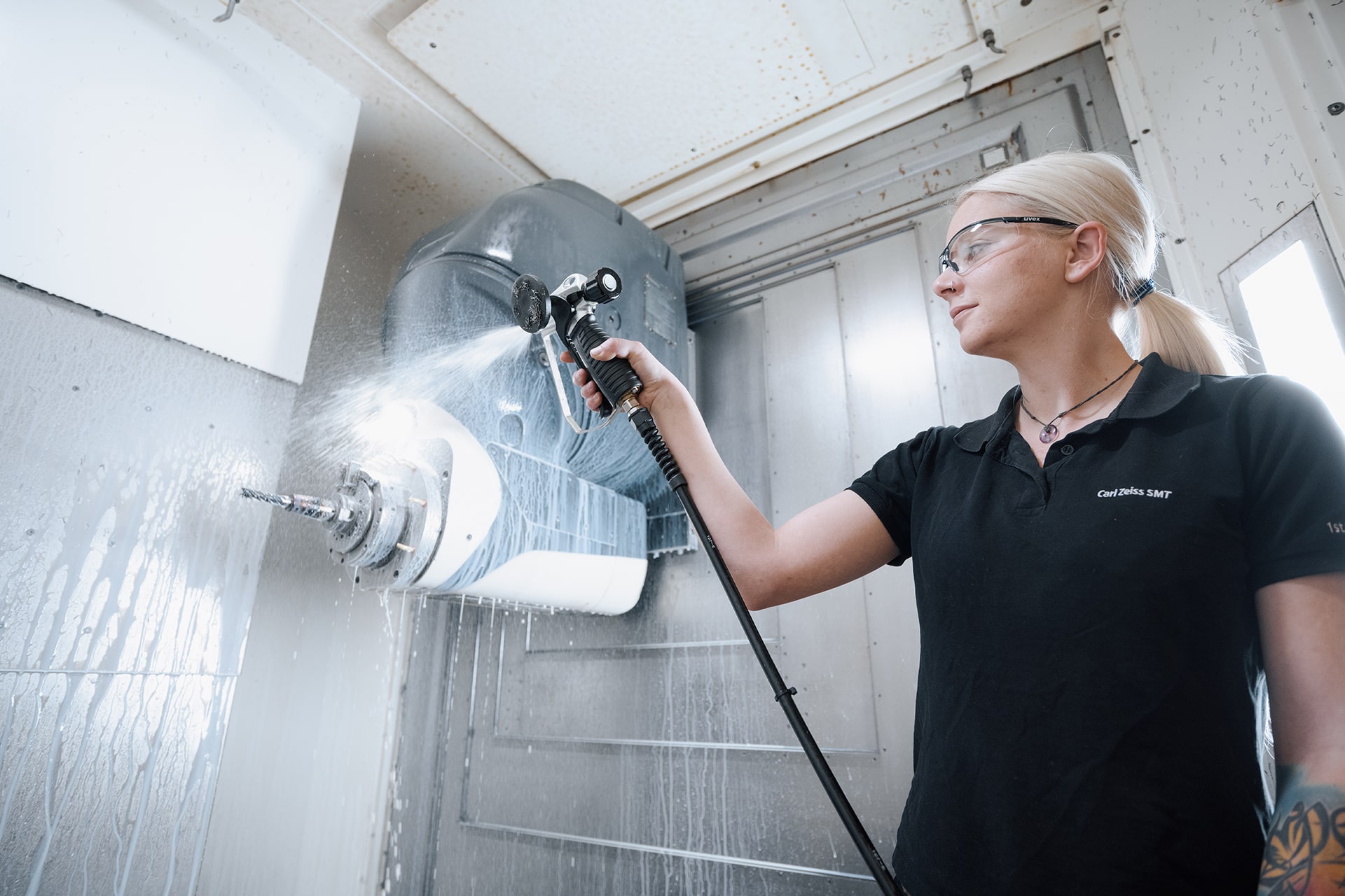 Melanie holds a water hose in her left hand and sprays a device. She is wearing safety goggles and a black T-shirt with &quot;Carl Zeiss SMT&quot; embroidered on it. The device is attached to a metal wall