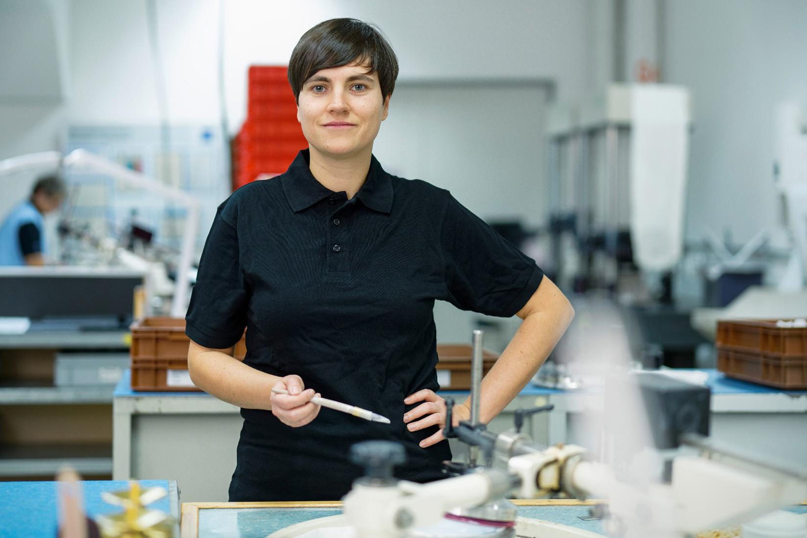 Sandra steht im Labor und schaut frontal in die Kamera. Sie hält einen Pinsel in ihrer rechten Hand fest und stützt ihre andere Hand auf ihrer Hüfte