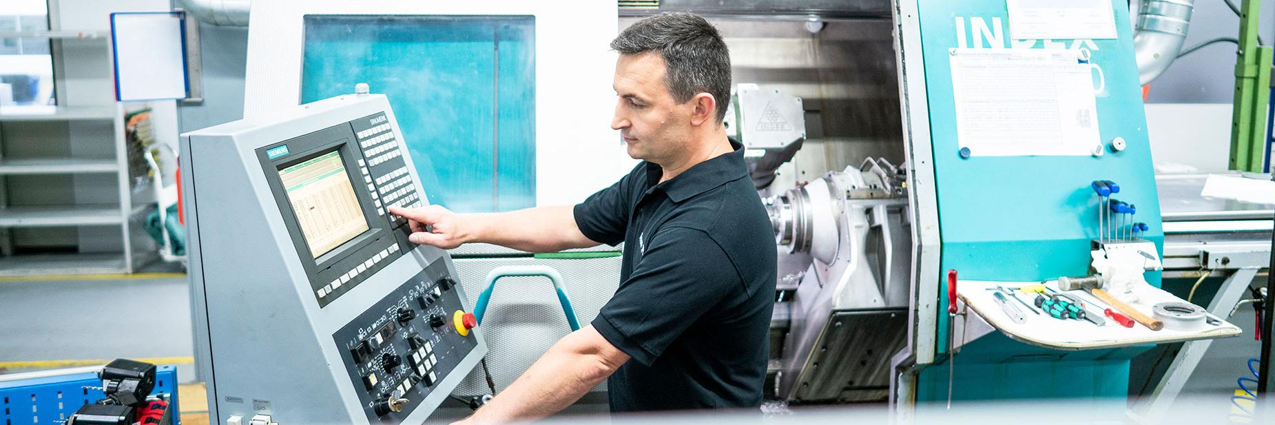 Waldemar steht neben einem Steuerungspult und bedient eine metallverarbeitenden Maschine.